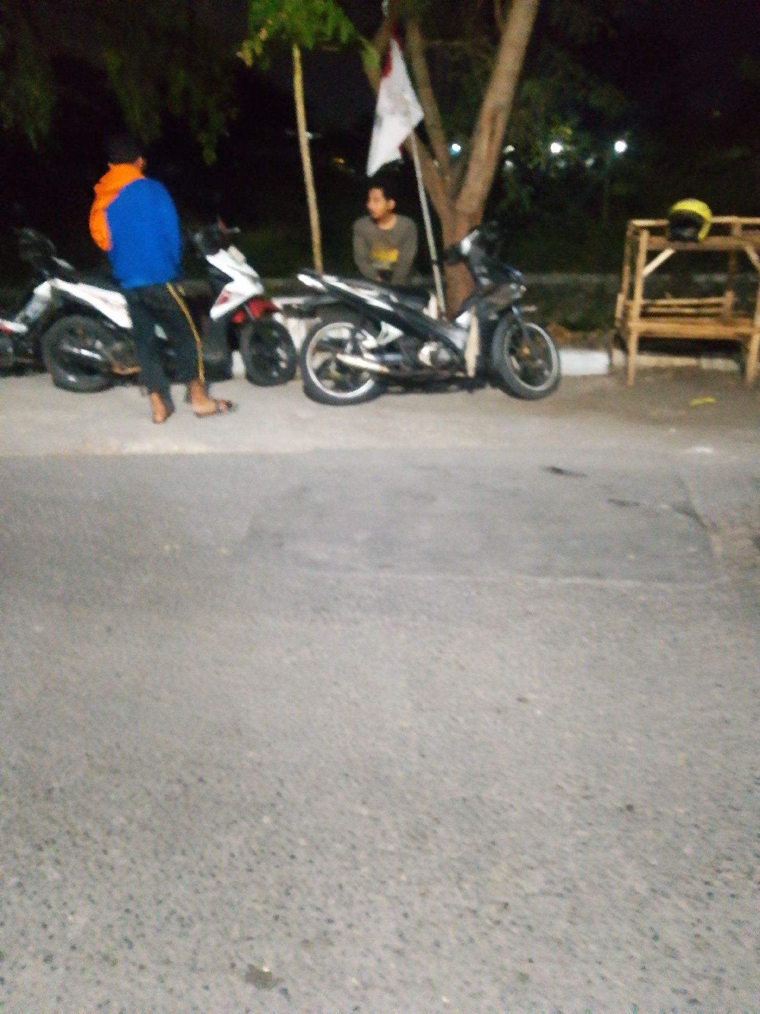 The image depicts an outdoor scene at night in Tambak Sumur, Indonesia. In the foreground, a paved road surface is visible, with a slight incline. In the midground, two motorcycles are parked. One motorcycle on the left is white and red, and the other on the right is black. A person wearing a blue jacket and orange accents is standing next to the white and red motorcycle. Another person, wearing a grey shirt, is seated on the black motorcycle. A wooden structure, possibly a bench, with a yellow helmet on top, is situated to the right of the motorcycles. A flag, predominantly white with a red stripe, is planted nearby. The background consists of trees and a dark, possibly skyward, expanse with some blurred lights.