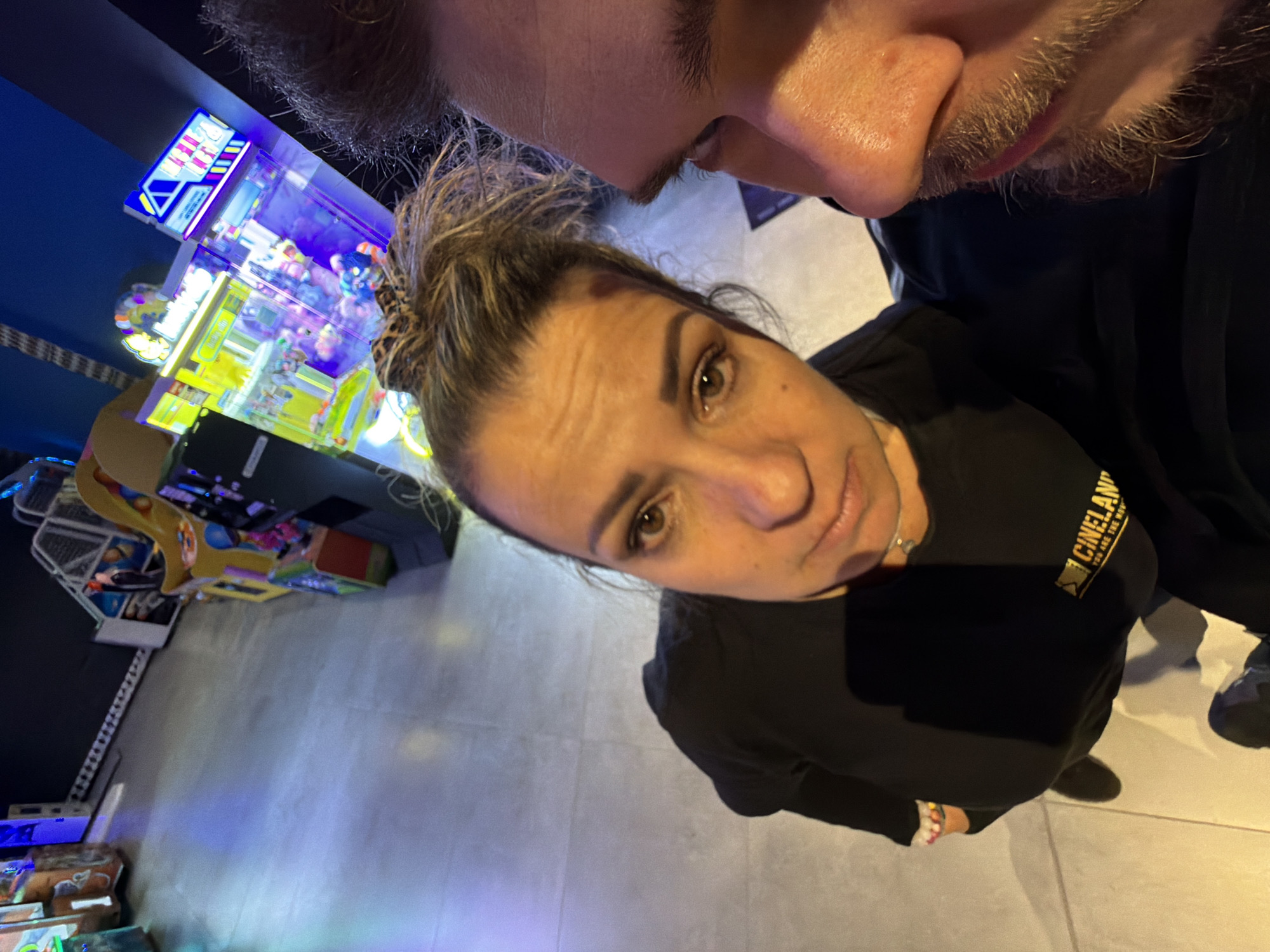 A close-up selfie shows two people inside what appears to be an entertainment arcade. The woman is in the center, looking at the camera. Her dark hair is in a bun. She wears a black t-shirt with "CINELAND" in gold lettering. The top of a man's face, including his nose and mustache, is visible above her. Arcade games, including a claw machine, are in the background. The floor is tiled. The photo was likely taken in Pernik, Bulgaria, as indicated by the CINELAND logo, which is a cinema chain with a location in that city.