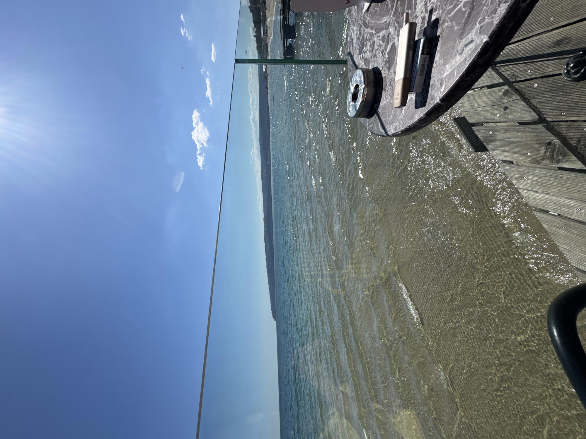 The image presents a seaside scene, likely taken in Varna, Bulgaria, given the provided context. A blue sky with scattered clouds is visible above the clear water. In the foreground, a round table with an ashtray, an electronic smoking device, and a pack of cigarettes sits on a wooden deck. A glass barrier separates the deck from the water, which appears shallow and clear near the shore. The water extends towards the horizon, where land is visible in the distance. A black railing curves into the frame at the bottom right, partly obstructing the view.
