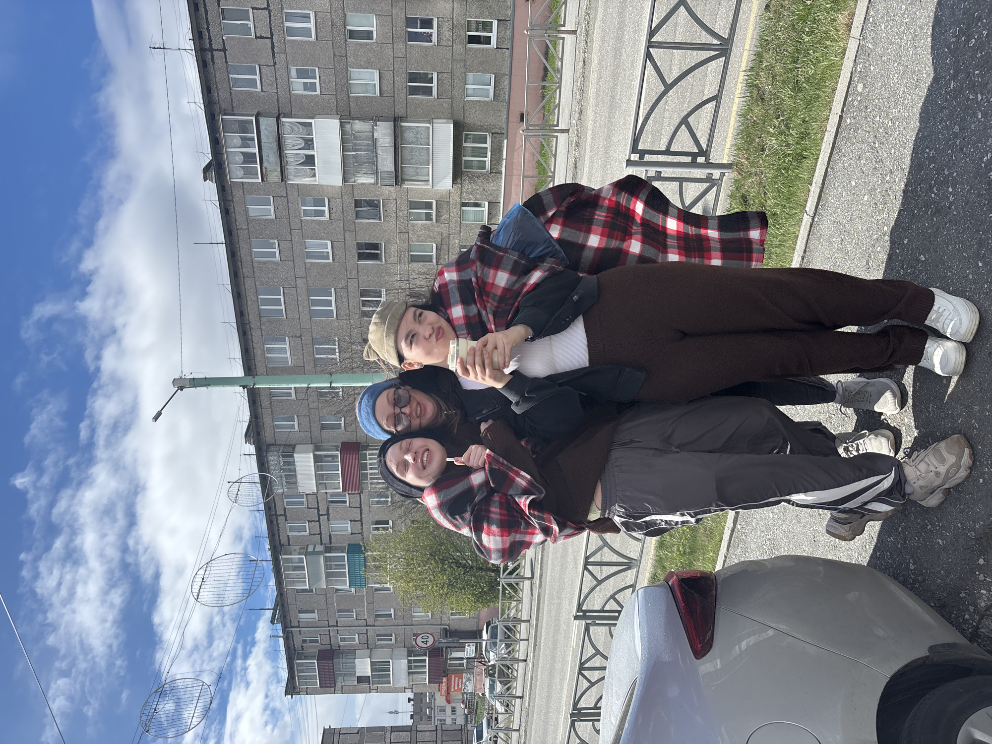 Three people are posing in an urban environment. The background consists of a gray, multi-story apartment building with multiple windows and possibly balconies. The sky is visible with patches of blue and white clouds. Power lines are present, decorated with circular decorative elements. A streetlight is also visible. The group is positioned near a curb and part of a car is visible at the bottom of the frame. The setting is identified as Kachkanar, Russia.