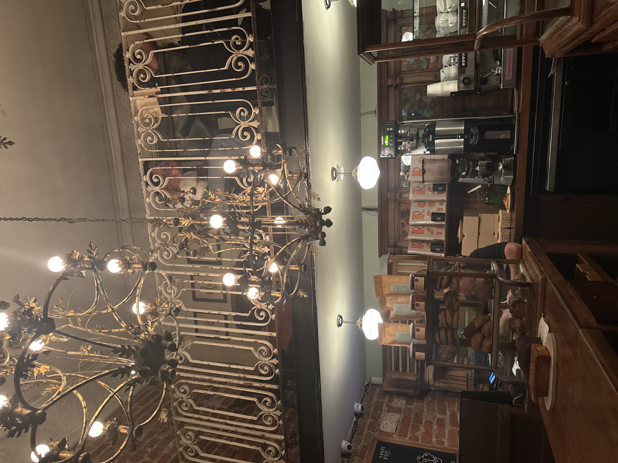The image shows the interior of a café or restaurant located in Mexico City, Mexico. The view is oriented upwards, showcasing architectural details and interior design elements.
In the foreground and left side is an ornate, multi-tiered chandelier with numerous light bulbs, suspended from the ceiling. The chandelier is predominantly gold and black.
Above the chandelier is a decorative wrought-iron railing. People are visible behind and between the railing, suggesting a mezzanine or upper level.
On the right side, a wooden counter or display case holds baked goods. Behind the counter, shelves are stocked with products, including coffee bags. A person is visible behind the counter, and coffee machines and other equipment are visible.
The walls of the establishment appear to be brick. Wall-mounted lighting fixtures are present.
The overall atmosphere appears to be dimly lit and upscale, typical of a historical or boutique establishment.