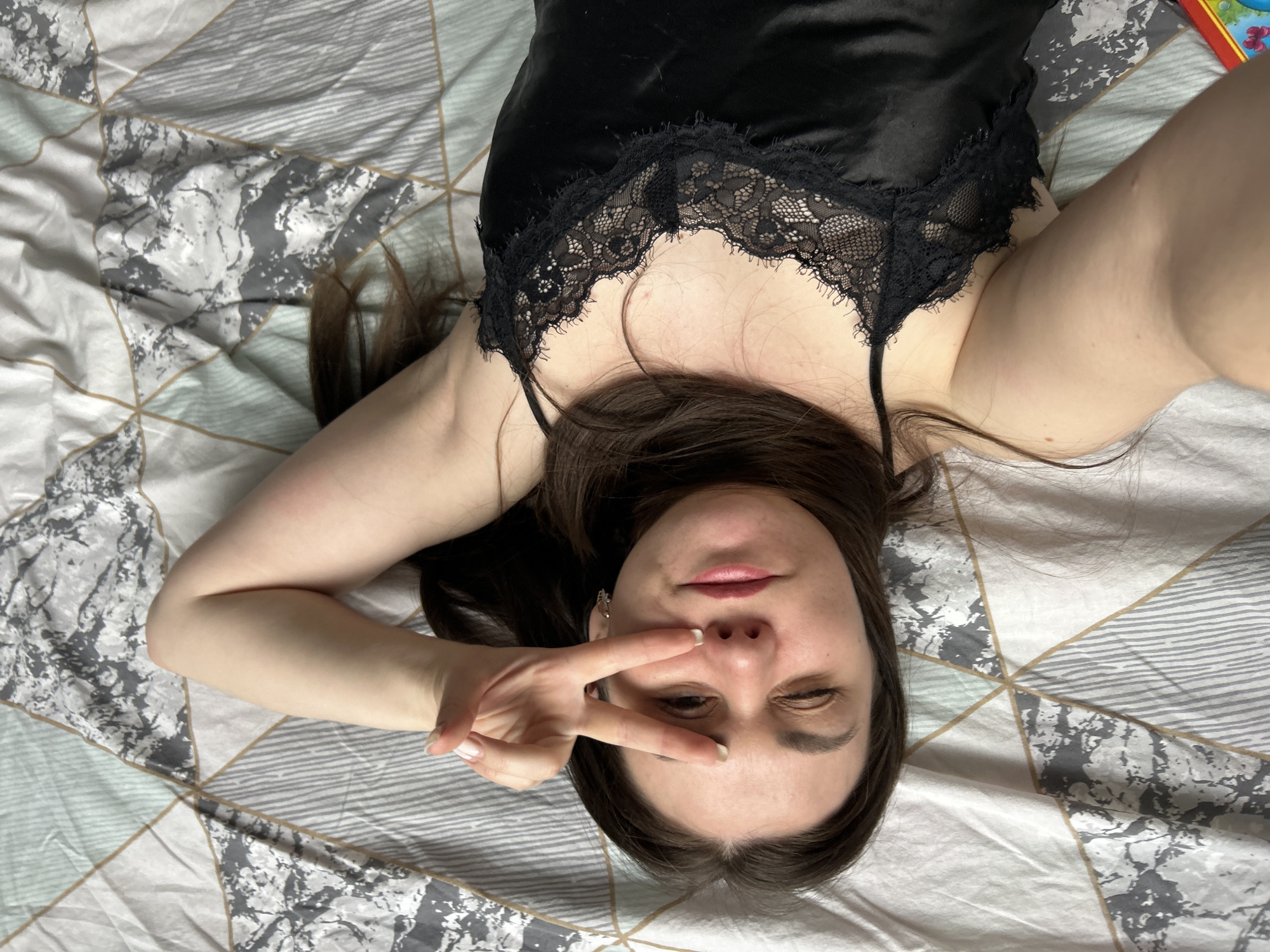 A young adult female is depicted in a close-up selfie, lying down on a bed and looking up at the camera. Her head is positioned towards the bottom of the frame, with her long, dark brown hair fanned out around her. She wears a black satin camisole or top, featuring a delicate black lace trim along the neckline.
The woman has a playful expression, with her left eye closed in a wink and her right eye partially open. Her lips are slightly parted. Her left hand is raised to her face, forming a "peace" sign (V-sign) with her index and middle fingers, while her thumb rests against the side of her nose.
She is lying on bedding covered by a duvet or blanket with a modern geometric pattern. The pattern consists of various sized triangles and squares in shades of white, light grey, dark grey, and a subtle light blue-green, outlined by thin light brown or gold lines. In the upper right corner of the image, a small, colorful object, possibly a book or toy, is partially visible. The scene is brightly lit, suggesting an indoor daytime setting. There is no visible text in the image.