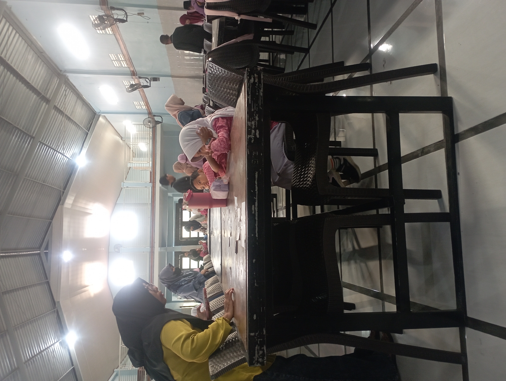 In Nganjuk, Indonesia, a group of people are gathered inside a well-lit room with a high, corrugated metal ceiling. The room appears to be a community hall or a large dining area. A long, dark wooden table dominates the foreground, with several chairs made of woven material surrounding it. Several individuals, including women and children, are seated or standing around the table. Some of the children are looking down at the table, possibly engaged in an activity or meal. A woman in a yellow shirt and black hijab stands to the left, looking towards the table. The floor is tiled with a reflective, light-colored surface, and the walls are painted a pale blue. Fans are mounted on the walls, indicating a warm climate. The overall atmosphere seems to be one of a casual gathering or event.