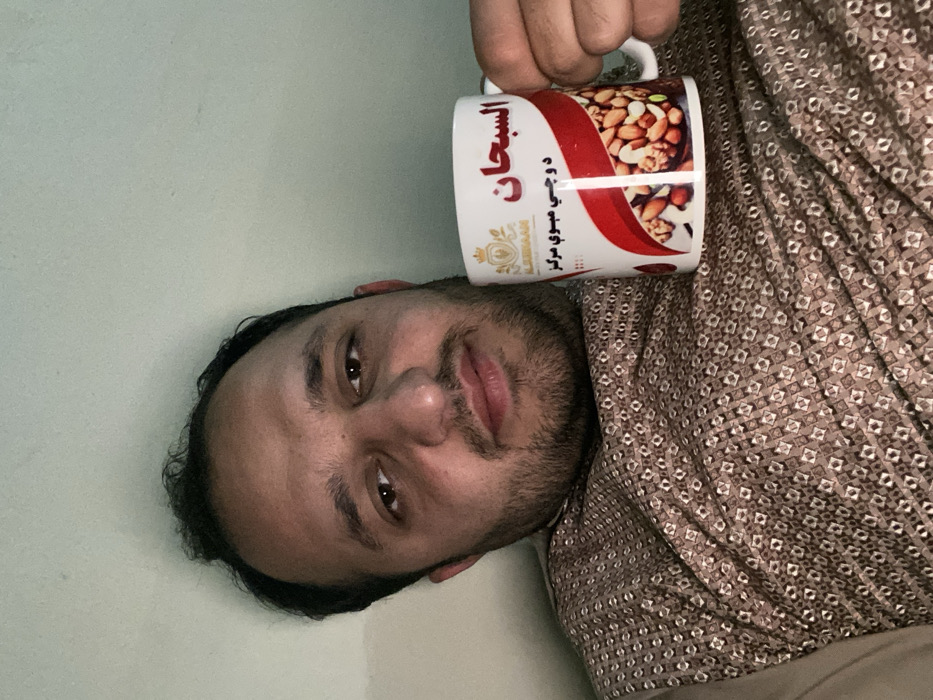 A man is holding a mug with nuts printed on it. The mug has Arabic text. He is wearing a patterned shirt with small square designs. The man has dark hair and a beard. The background appears to be a plain wall. The image is oriented sideways.