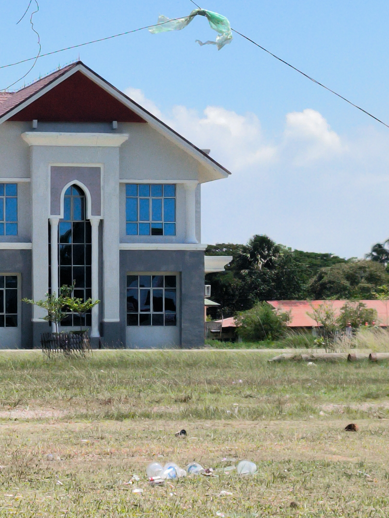 The image depicts a two-story building with a red-brown roof in Meunasahaluedrienlhoksukon Barat, Indonesia. The building has light gray exterior walls with gray accents around the windows and the central, arched window on the second floor. The windows are blue and reflect the sky. A small tree is planted in front of the building, enclosed by a small fence.
The foreground is an overgrown grassy field littered with plastic bottles and other trash.
In the background, beyond the building, are trees and another structure with a red roof. A piece of green plastic is caught on a high wire above the building.