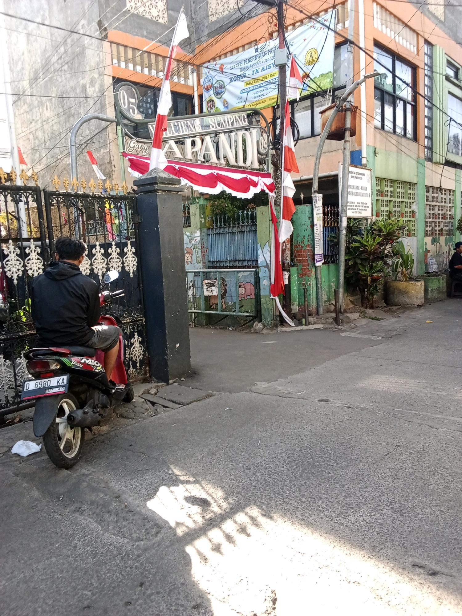 The image depicts the exterior of a building identified as TK AISYIYAH BUSTANUL ATHFAL 05, located in Bandung City, Indonesia. The building has a facade with orange and green sections, and multiple windows. Red and white Indonesian flags are prominently displayed, both on poles and as decorative banners. A black metal gate with ornate detailing is visible in the foreground.
A person is seated on a red and black motorcycle, facing away from the camera, positioned near the gate. The motorcycle's license plate reads "D 6980 KA". The ground is paved with asphalt, and there are some cracks and debris visible. The scene appears to be a street in front of the educational institution, with shadows cast on the pavement, suggesting it is daytime. To the right of the building, a person is seen sitting on a stool near a wall. The overall impression is a snapshot of a typical street scene in an urban Indonesian setting, likely in the morning or afternoon due to the sun's position.