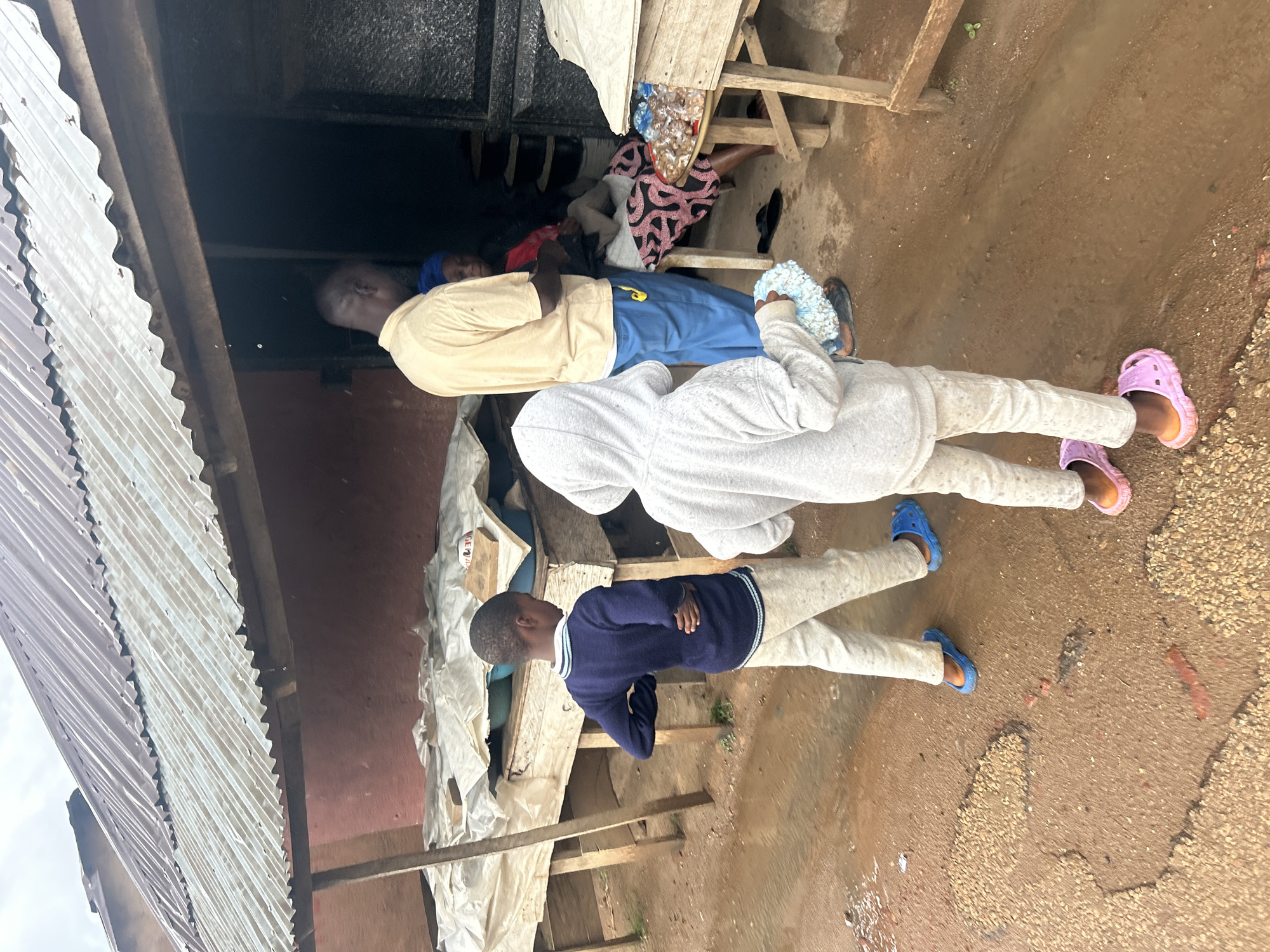 This media file presents an outdoor scene, likely in Jos, Nigeria, featuring four individuals amidst makeshift structures. The image is oriented with the ground on the right and structures on the left.
In the center-right, an adult male stands upright, wearing a yellow short-sleeved shirt and blue trousers, with his head turned towards the top of the frame. Partially obscured behind him, another individual is seated, dressed in patterned fabric. To the male's left, a person in a grey hooded sweatshirt and light-colored trousers stands, also facing upwards, wearing light blue and white fluffy slippers. Further to the left and slightly below, a child in a navy blue sweater with light blue stripes and light-colored trousers stands with crossed arms, looking towards the left. This child wears blue plastic clogs.
The ground, prominent on the right, is dirt, appearing damp and uneven with patches of small stones. On the left, a corrugated metal roof angles across the frame. Behind the people, various wooden planks form a makeshift stall or shelter, laden with items like plastic bottles and fabrics. A dark, possibly metal, doorway or gate is visible in the background. The sky, seen briefly at the far top-left, appears overcast, suggesting daytime. The scene depicts everyday activity in what appears to be an informal settlement or market environment. Visible text "SE" can be discerned on a white tarp on the far left side.