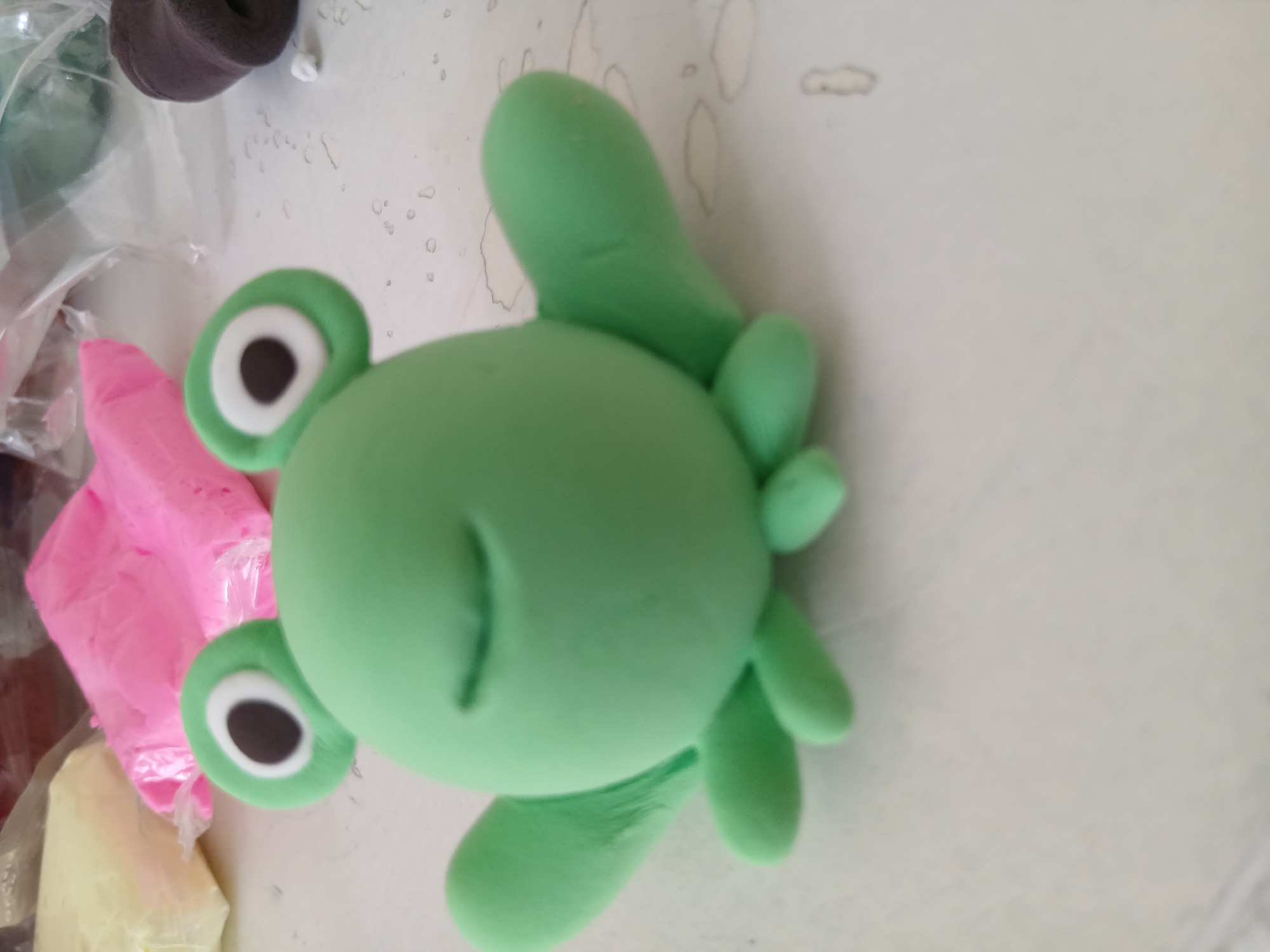 The image shows a bright green, cartoon-like figure, possibly a frog or turtle, made from modeling clay or play-doh. The figure is positioned horizontally and is the main subject. It features two large, round eyes with white irises and black pupils, surrounded by green, and a simple, curved mouth line. Several rounded, stubby limbs or flippers extend from its body.
In the background, other crafting materials are visible. To the left of the green figure, there is a block of vibrant pink modeling clay partially wrapped in a plastic bag. Below it, a yellowish block of material, also likely clay, is present. In the upper left corner, a dark brown or black irregularly shaped object, possibly more modeling clay, can be seen. All the items are resting on a light-colored, flat surface, which has some faint smudges and outlines, suggesting a crafting or play area. The scene depicts the result of a creative activity involving modeling with clay. There is no visible text.