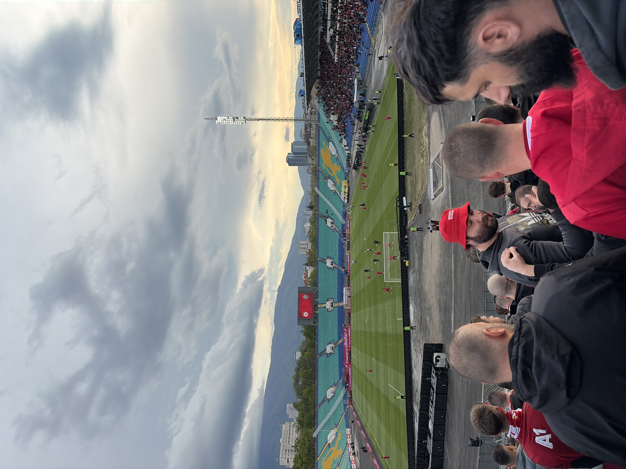The image captures a vibrant football stadium scene in Sofia, Bulgaria, likely during a late afternoon or early evening match. The sky is partially cloudy with patches of warm, golden light breaking through, suggesting a transitional time of day.
In the immediate foreground, several male spectators are visible from behind or in profile. One man wears a red beanie with "LUX" visible text, another has a prominent beard and is wearing a red jacket, and others are in dark hoodies or jackets, including one with an "adidas" logo. Their attention is directed towards the field.
The midground features the football pitch, where players in red and white kits are active, indicating a match is underway. The stadium stands are packed with numerous spectators, with distinct sections of blue and green seating. A large scoreboard or screen displays a red background with a white and red crest or logo, and "OMV" is visible on an advertising board beneath it. Tall floodlight towers illuminate the field, with one particularly prominent on the left. Advertising hoardings along the pitch side display text like "Powered by ultrafan.tv".
In the background, a cityscape with various buildings is visible, set against the backdrop of imposing mountains, which are characteristic of the Vitosha Mountain range near Sofia. The overall atmosphere is that of a live sporting event enjoyed by a significant crowd.