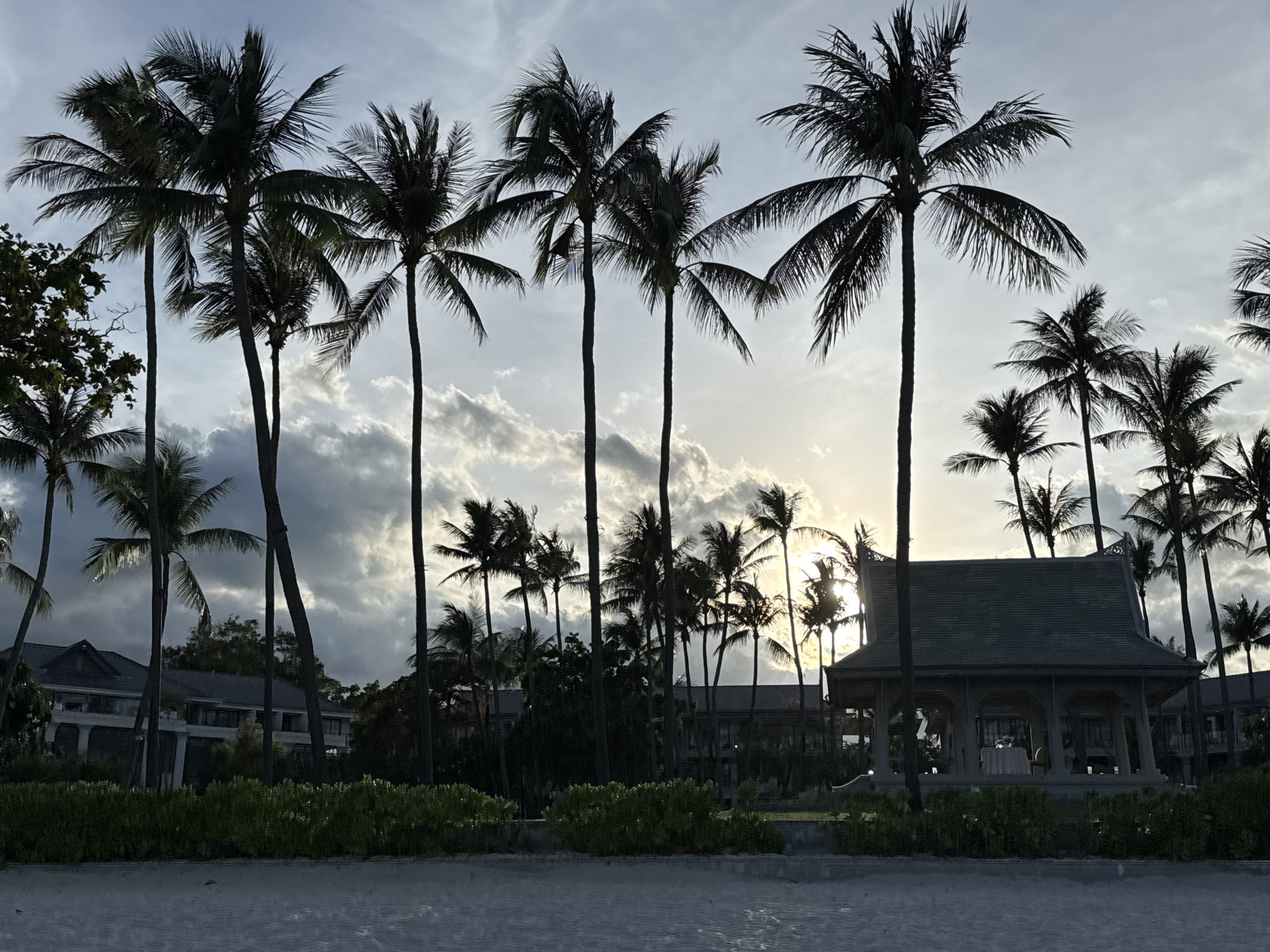 A wide shot of a beach scene in Baan Chaweng, Thailand, shows silhouetted palm trees against a cloudy sky with the sun peeking through. The background reveals structures that appear to be buildings, possibly part of a resort. In the lower right, a pavilion-like structure with a table and chairs inside stands. Low green shrubbery lines the front of the scene, with sand visible at the very bottom. The overall scene suggests a tropical setting at dusk or dawn. No people are visible.