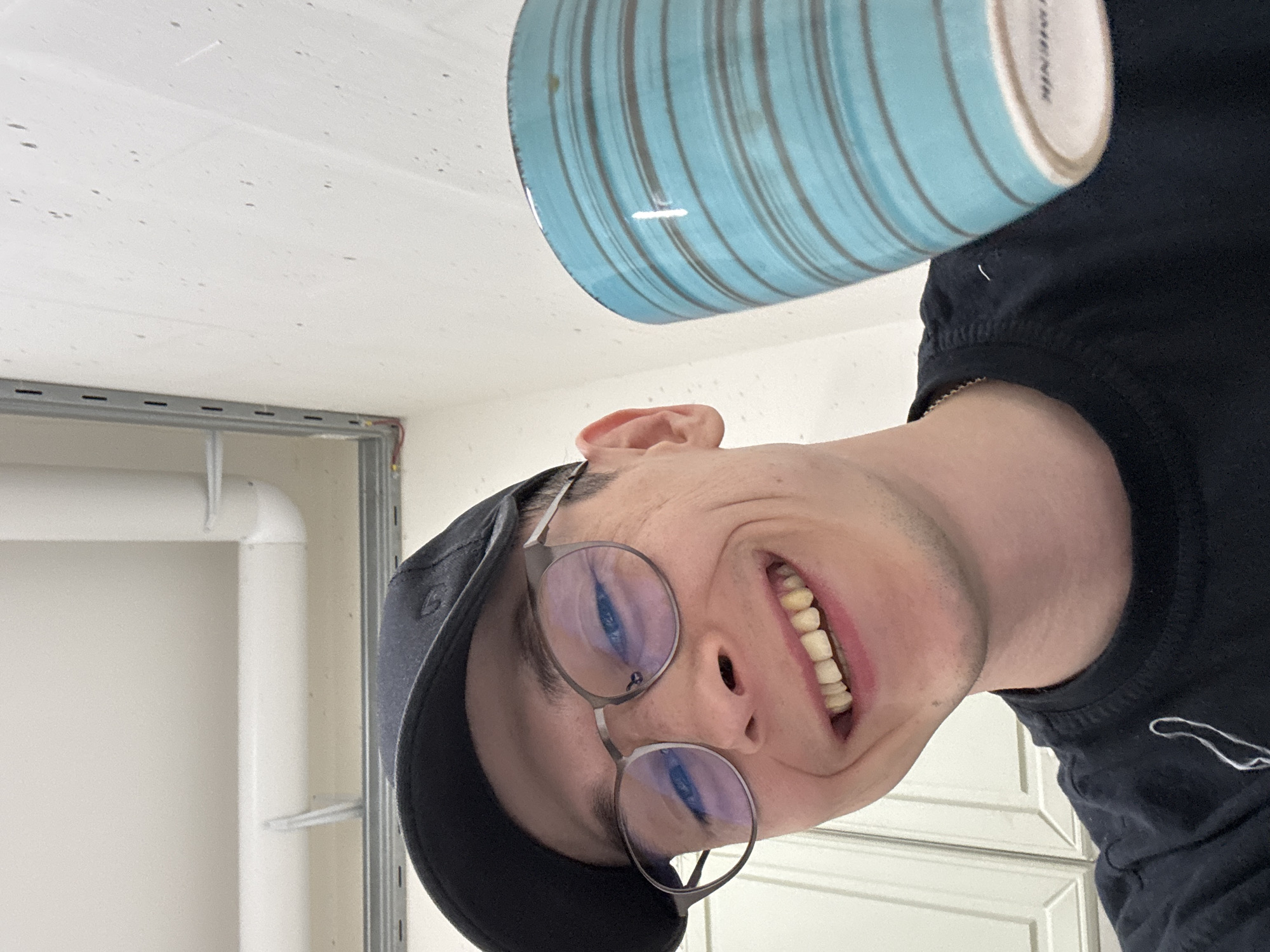 A young Caucasian male wearing glasses and a black baseball cap is holding a blue striped mug. He is smiling broadly, showing his teeth. The background appears to be indoors, possibly a kitchen or storage area, with white cabinets visible. There are exposed pipes and wiring on the ceiling, suggesting a utilitarian or unfinished space. The lighting is bright. The provided image crops do not offer any specific details about the city of Ostrov, Russia, or any landmarks that would indicate this location.
