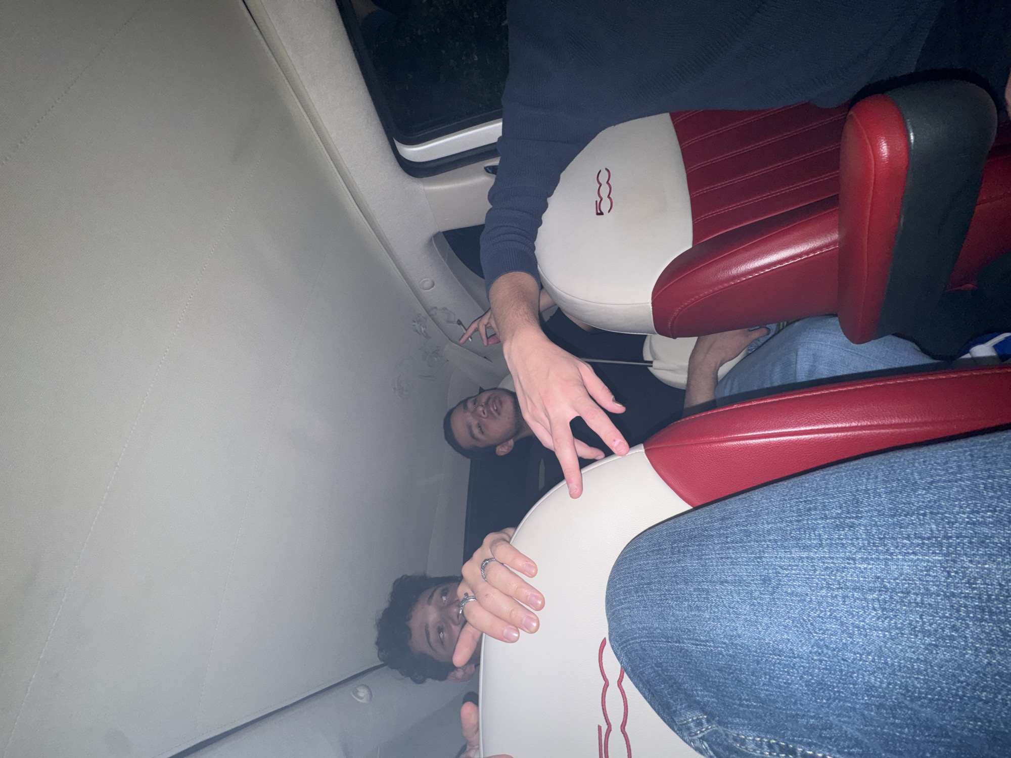 The image appears to be taken inside a car, potentially a Fiat 500, judging by the seat design. There are two people visible. One is looking directly at the camera, with a curly dark hair and wearing a ring on his fingers. The second person is in the upper center, also looking towards the camera, and has their hand stretched out. A third person may be present as indicated by the arm. The interior of the car is light-colored with red and white seats. There is a sunroof visible. It's impossible to determine if this is Mexico City without additional information about the location.