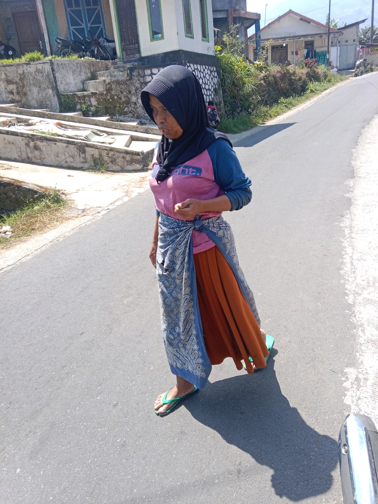 A medium shot shows a woman walking on an asphalt road in the city of Adipuro, Indonesia. She is wearing a black hijab, a pink and blue long-sleeved shirt, and an orange skirt with a blue patterned sarong tied around her waist. Her feet are clad in green flip-flops. The woman's head is turned to her left, and her right hand is extended slightly forward as if holding something. Her shadow is cast on the road to her right. In the background, a building with a blue door and windows is visible to the left, and houses with tiled roofs are visible to the right. There are also motorcycles parked near the building. The environment appears to be a rural or semi-urban area with buildings and vegetation. The lighting suggests it is daytime, with a clear sky.