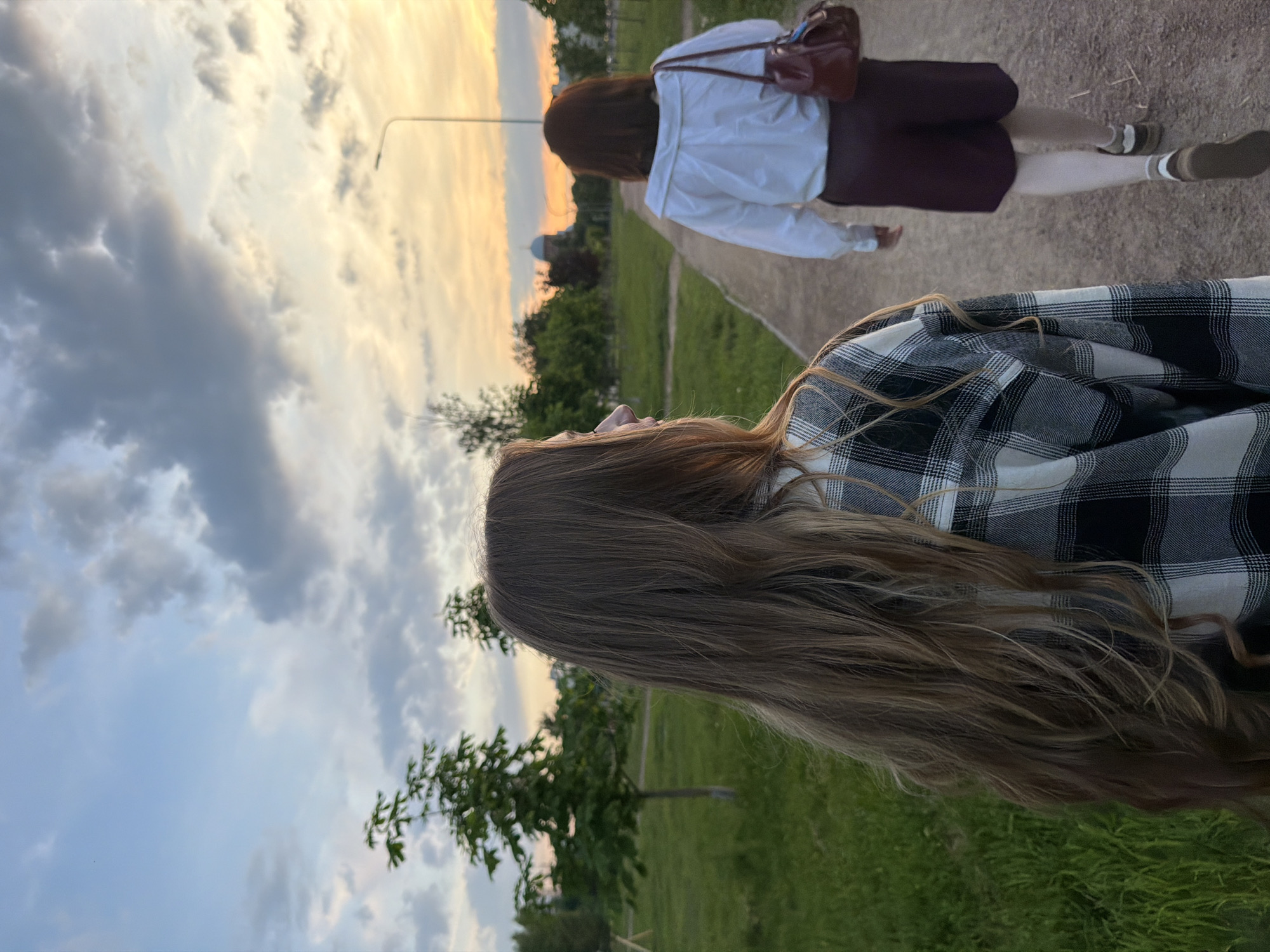 Two individuals are walking away from the camera on a paved pathway in a park-like setting during sunset. The person in the foreground, on the left, has long, light brown hair and is wearing a black and white plaid shirt. The person in the background, on the right, has dark brown hair and is wearing a white collared shirt and dark red shorts, carrying a burgundy handbag over their right shoulder. The pathway is bordered by green grass and scattered trees. In the distance, a building with a dome is visible, along with streetlights and more trees, suggesting an urban or suburban park environment. The sky displays a gradient of colors typical of sunset, with clouds and a warm glow. The location is identified as Sinyavino, Russia.