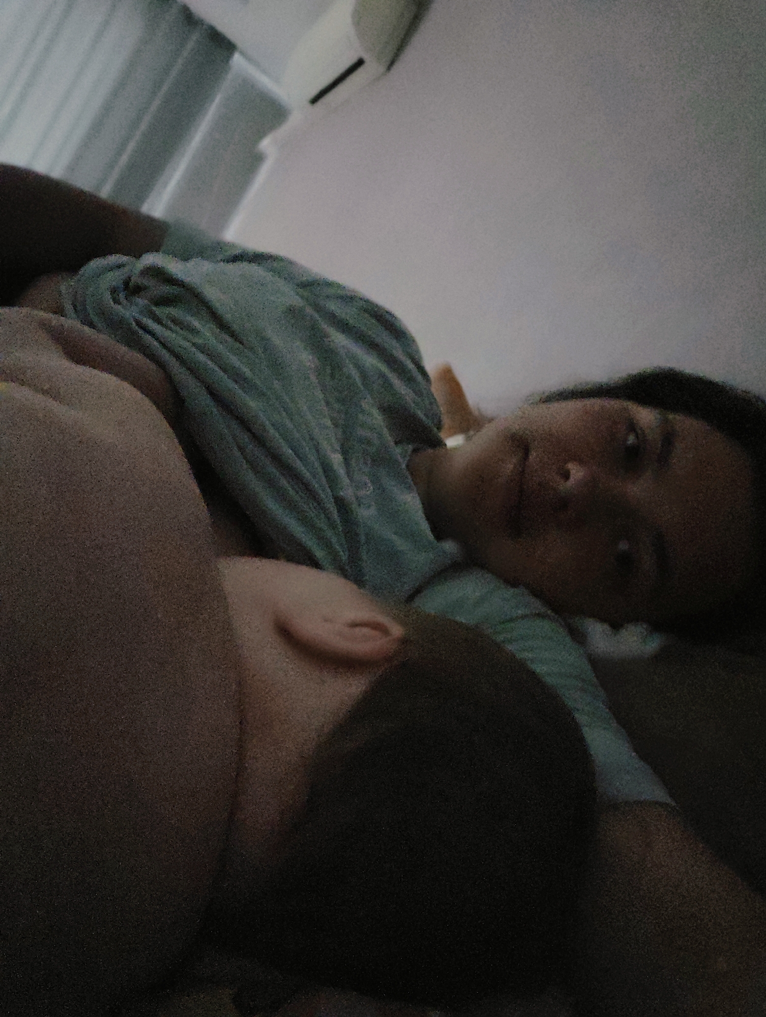 A woman and a baby are lying in a bed indoors. The woman is lying on her back with her head turned towards the camera, and her face is visible. She has dark hair and is wearing a green t-shirt. A baby is lying on its side next to her. The baby's head is visible, and it has dark hair and a small ear. The background shows a wall and curtains. The location is unknown.
