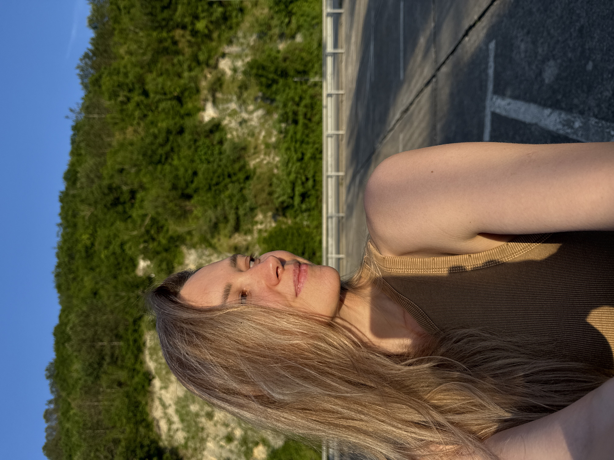 This image depicts a young adult female outdoors in Nebug, Russia, under clear, sunny conditions. The female, with long, light brown hair and light skin, is presented from the upper torso, turning her head towards the right side of the frame with a slight smile. She is wearing a ribbed, tan-colored sleeveless top. Sunlight illuminates her left side, casting shadows on her right. The background features a grey asphalt surface with faded white markings, horizontally traversed by a metallic guardrail. Beyond this, a steep hillside covered with dense green vegetation and sections of light-colored earth rises into a clear blue sky.