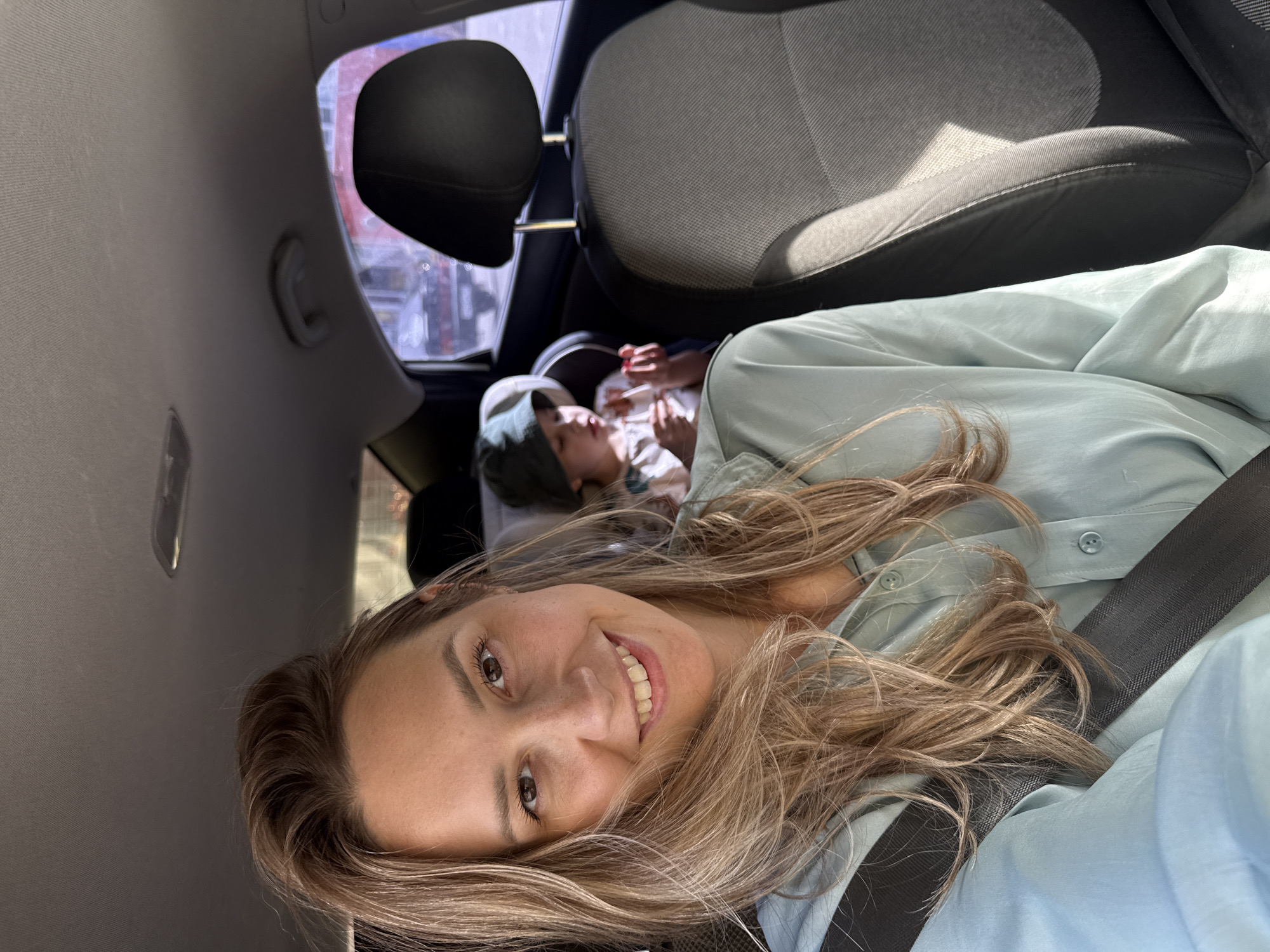 The image shows a woman in the front seat of a car, wearing a seatbelt and light blue shirt, smiling towards the camera. In the background, a child is sleeping in a car seat in the back. Outside the car window, blurry buildings are visible, suggesting an urban environment. The image location information suggests that the photo may have been taken in Chaikovsky, Russia.