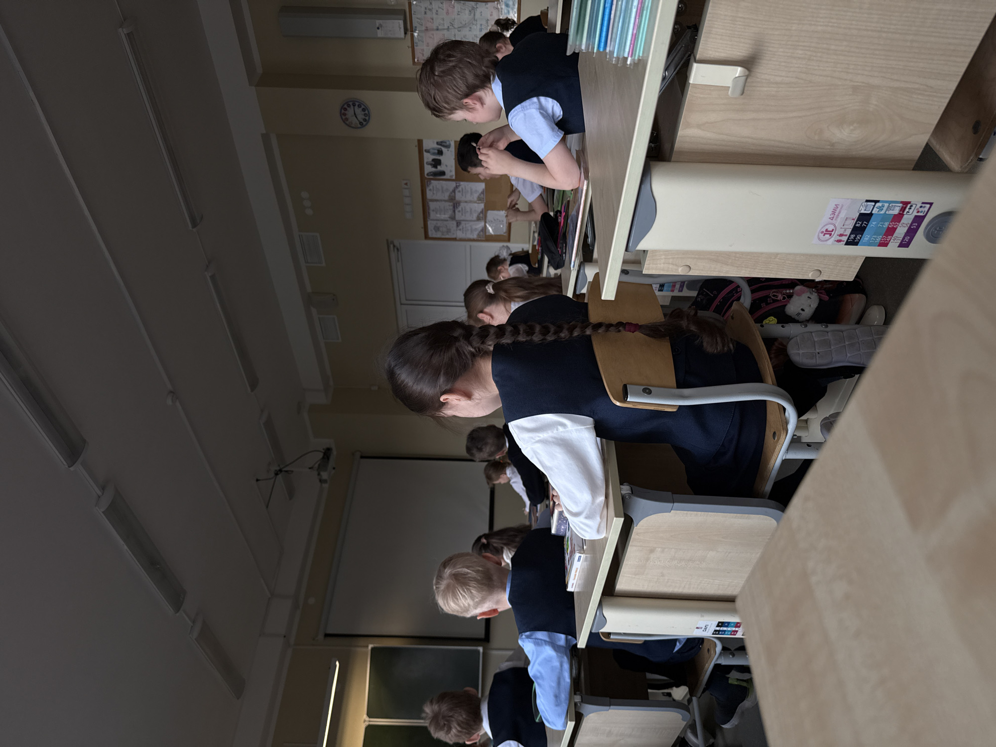 The image captures a classroom scene, viewed from a slightly elevated position towards the front. Several students, primarily children, are seated at individual desks arranged in rows. They are dressed in school uniforms, consisting of dark vests or jumpers worn over light-colored shirts or blouses. Most students are seen from behind or from a side angle, with their heads bent forward, suggesting they are focused on tasks at their desks, possibly reading or writing.
The classroom features typical school furniture: light-colored wooden desks and chairs with metal frames. Overhead, fluorescent light fixtures illuminate the room, and a large projector screen or whiteboard is visible at the front. A circular wall clock with Roman numerals is present on the wall in the background, though the exact time is difficult to discern. Underneath one of the desks, various personal items like backpacks, shoes, and a small white plush toy are visible.
A notable detail is a sticker on the side of one of the desks, indicating it is adjustable. The sticker features the text "ДЕЛИ" (DELI) and a table with height measurements in centimeters (e.g., 198, 185, 173, 160, 146, 134, 120) correlated with numerical or color codes, suggesting different settings for various student heights. The overall impression is of an ordinary school day, with students engaged in learning in a well-equipped classroom in Perm, Russia.
