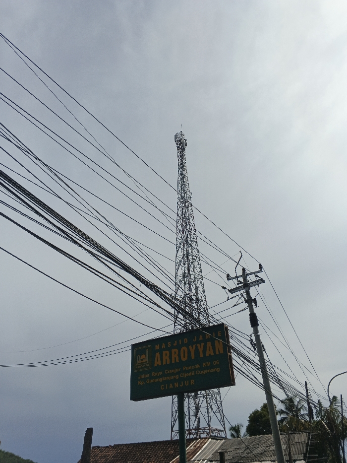 The image features a communication tower against a pale sky, cluttered with numerous overhead power lines. A green sign labeled "MASJID JAINE ARROYYAN" stands in the foreground, providing directions to Jalan Raya Cianjur Puncak KM 06, Kp. Gununglanjung Cijedil Cugenang, Cianjur. A utility pole with transformers is positioned to the right of the sign. The base of the tower and the sign are partly obscured by rooftops of nearby buildings.