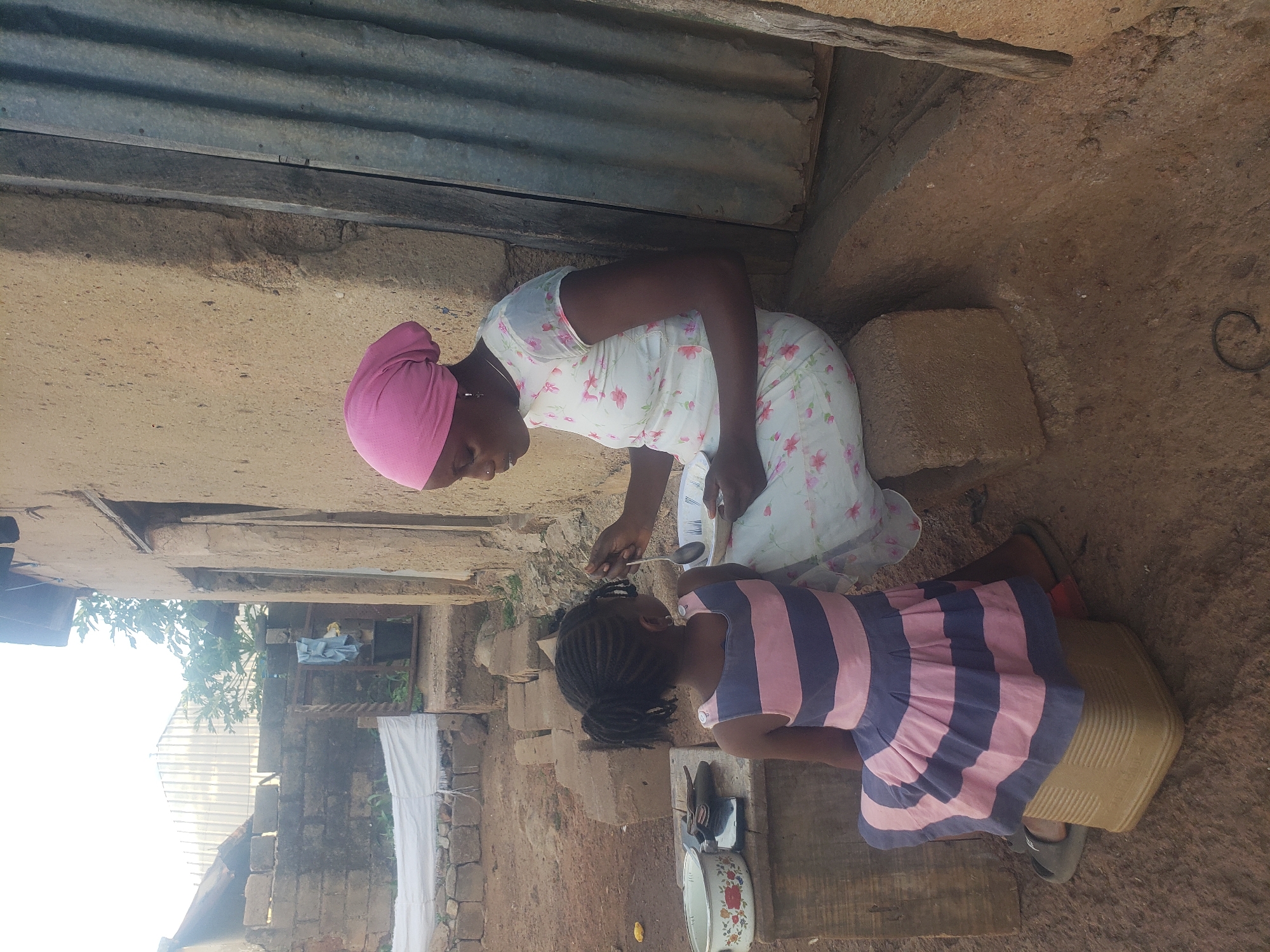 A woman wearing a pink headscarf and a white floral dress is kneeling on a stone surface, feeding a girl in a striped dress with a spoon. The girl is seated on a beige plastic container. Cooking pots and other utensils are on the ground nearby. The setting appears to be outdoors in an urban area, with building materials and structures visible in the background. The location is identified as Bukuru, Nigeria.