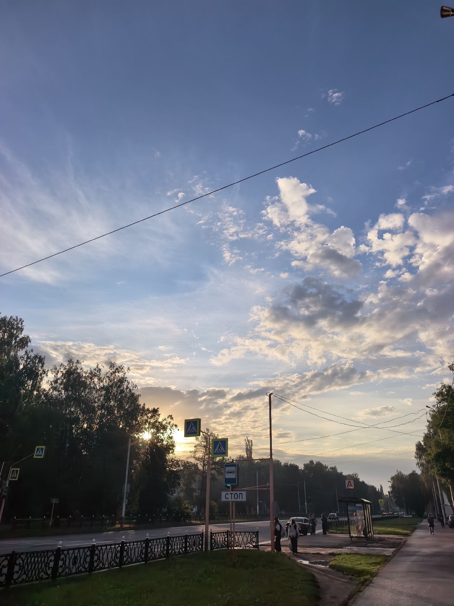This image depicts a street scene in Sterlitamak, Russia, at dusk. The sky is a gradient of blue and white, with wispy clouds and a hint of sunset glow peeking through the trees on the left. A power line stretches across the upper portion of the frame.
The foreground features a sidewalk and a road. A decorative black metal fence separates a grassy area from the road. On the sidewalk, there's a bus stop shelter with a few people standing inside and nearby. Several pedestrians are also visible walking along the sidewalk and crossing the street at a marked crosswalk. Traffic signs, including pedestrian crossing signs and a "STOP" sign in Russian, are present. Several cars are parked along the road and one is in motion. Trees line the street on both sides, with dense foliage on the left and sparser trees on the right. The overall impression is a typical urban street scene transitioning from day to evening.