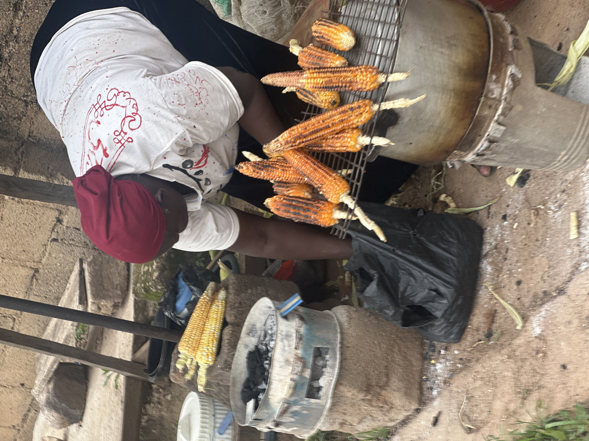 A woman wearing a red headscarf and a white t-shirt adorned with red paint splatters and decorative red markings is roasting corn on a grill in Gura Top, Nigeria. The corn is arranged on a metal grate over a large metal pot, which is itself placed on a rustic stove. Several cobs of corn are visible, some partially cooked, displaying charred kernels. In the background, unroasted corn rests on stones, and a black plastic bag lies on the ground. A charcoal stove, used for heating, is also visible, positioned on a stone base. The ground is a mixture of dirt and scattered debris.