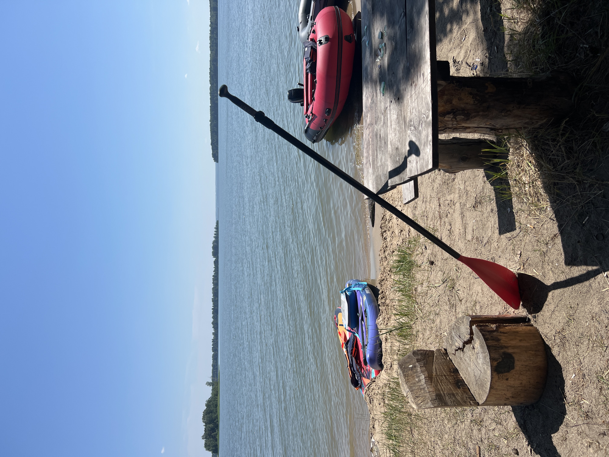 The image depicts a serene lakeside scene in Borovoe, Russia. In the foreground, a rustic wooden table made of logs sits on a sandy shore, casting a long shadow. A red-bladed paddle, leaning against the table, suggests recent activity or preparation for water sports. To the left, a unique chair carved from a tree stump stands near the water's edge.
The sandy beach is dotted with patches of dry grass. Several inflatable boats and paddleboards are partially submerged or resting on the sand near the water. A vibrant red inflatable boat with an outboard motor is moored in the shallows, alongside another dark-colored boat.
The calm, blue water of the lake stretches towards the horizon, where a dense line of green trees forms the distant shore. The sky above is a clear, bright blue, indicating fair weather. The overall atmosphere is peaceful and idyllic, suggesting a recreational spot by the lake.