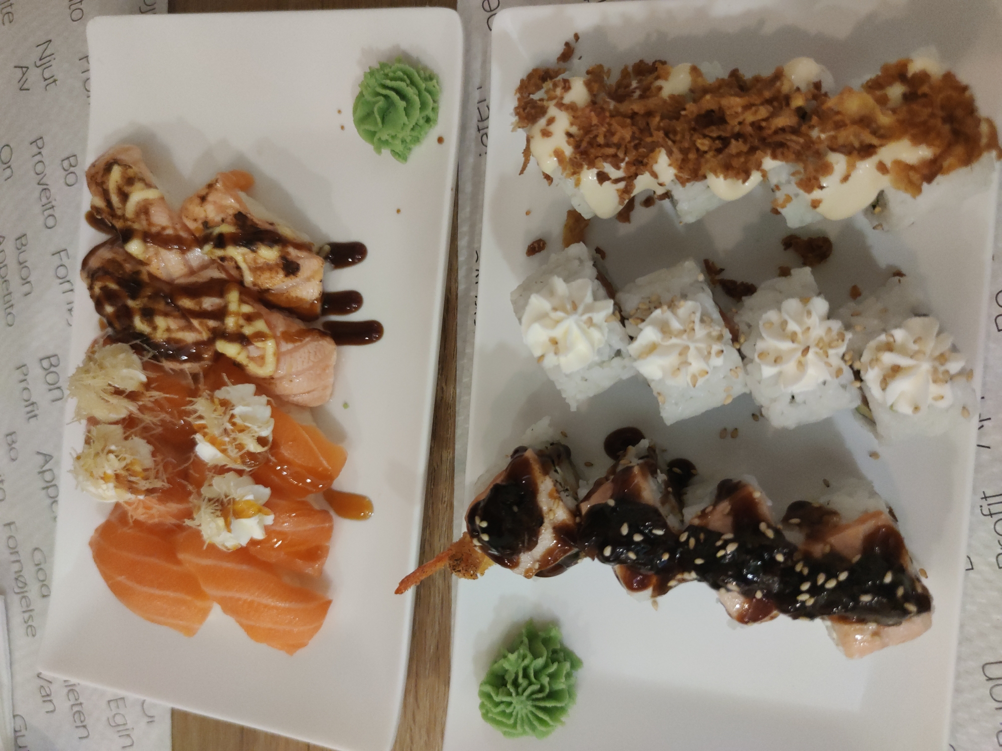 A top-down shot presents two white rectangular plates laden with various types of sushi, accompanied by small portions of wasabi on each plate. The left plate features an assortment of salmon sashimi topped with decorative elements and sauces, while the right plate contains sushi rolls, some adorned with a creamy topping and fried onions, and others with a dark glaze. The plates are positioned on a surface covered with a patterned paper.