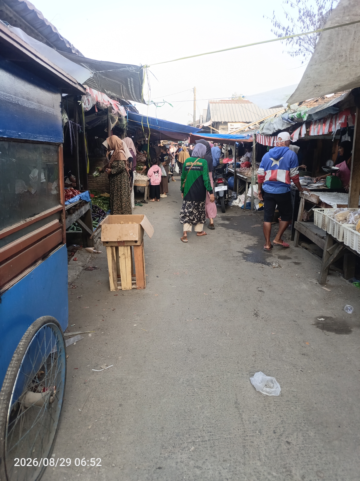 The image depicts a bustling outdoor market scene in Sangiang, Indonesia. The market is lined with stalls covered by awnings, showcasing various goods. In the foreground on the left, a blue cart with a spoked wheel is partially visible, suggesting a vendor's setup. The central path of the market is paved and shows signs of wear.
Several individuals are present, engaged in market activities. On the left, two women are seen near a stall with produce, one wearing a hijab and a patterned dress, and the other in a beige hijab. Further down the path, a person in a green jacket with white lettering and a patterned skirt walks away from the camera. To the right, a man wearing a blue hoodie with a star design and a white cap is facing a stall where fish are being displayed on a counter, with a scale visible. Another person in a black hijab is behind the counter. Children are also present; one in a pink jacket is visible near a stall.
The stalls display a variety of items, including vegetables like tomatoes and cucumbers, and fresh fish. The overall atmosphere suggests a typical day at a local Indonesian market, with the activities of buying and selling in full swing. The time stamp at the bottom of the image indicates August 29, 2026, at 06:52 AM.