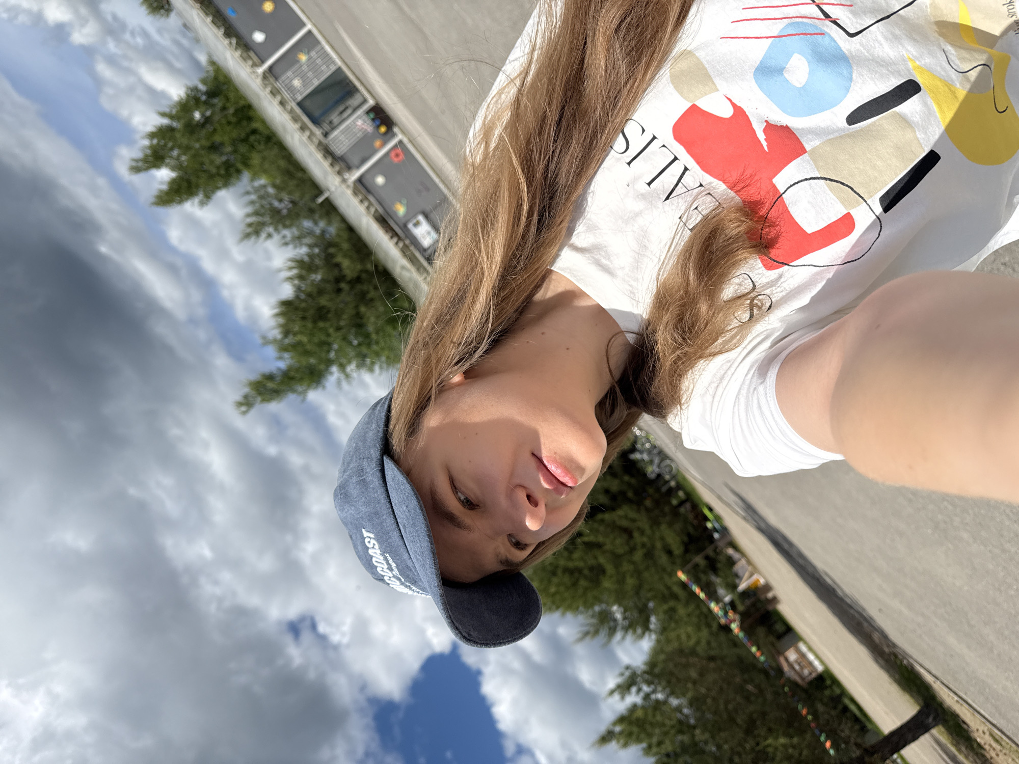 A young woman captures a diagonal selfie outdoors in Dobrograd, Russia. She has long, light brown hair and gazes towards the camera with a calm, neutral expression, her lips slightly parted. She is dressed in a white t-shirt adorned with a colorful, abstract design featuring red, blue, yellow, and black geometric shapes and lines. Visible text on her shirt includes "ALIS" and "Srok." She also wears a navy blue baseball cap with "COAST" embroidered in white on the front. Bright daylight illuminates the scene, casting shadows on her shirt and the paved ground below. The background features a partly cloudy sky, visible in the upper portion of the image. To her left, a grey, multi-paneled structure, possibly an informational board or utility unit, is visible. Trees and a string of colorful decorative lights or flags extend across the lower right background. The image depicts a daytime scene under fair weather conditions.