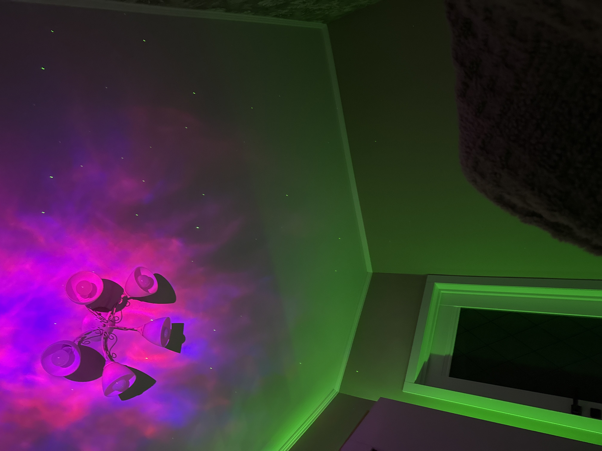 A low-angle view of a room features a ceiling decorated with a violet and magenta nebula-like projection, dotted with small, faint green lights resembling stars. A chandelier with multiple spotlights is mounted on the ceiling within the nebula projection. The walls are painted in a dark green hue. A section of a window frame is visible on the right side of the image. A large object, textured and dark in color, occupies the upper right corner, appearing as if it's partially obscuring the view.