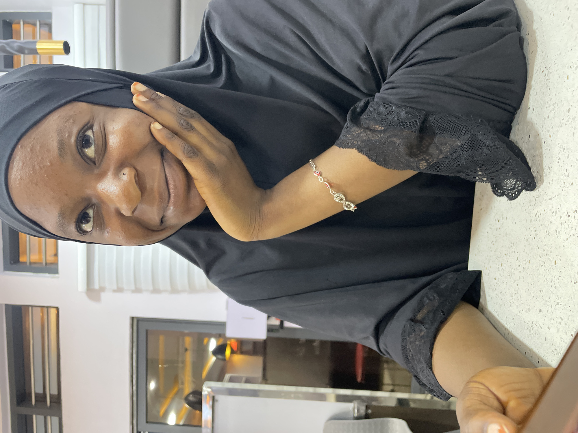 A woman wearing a black headscarf and black garment with lace trim on the sleeves is sitting at a white speckled countertop, presumably in a restaurant or cafe. Her left hand is gently touching her face. A silver bracelet with red and white details adorns her left wrist. The environment includes windows with security bars. The location is potentially Jos, Nigeria.