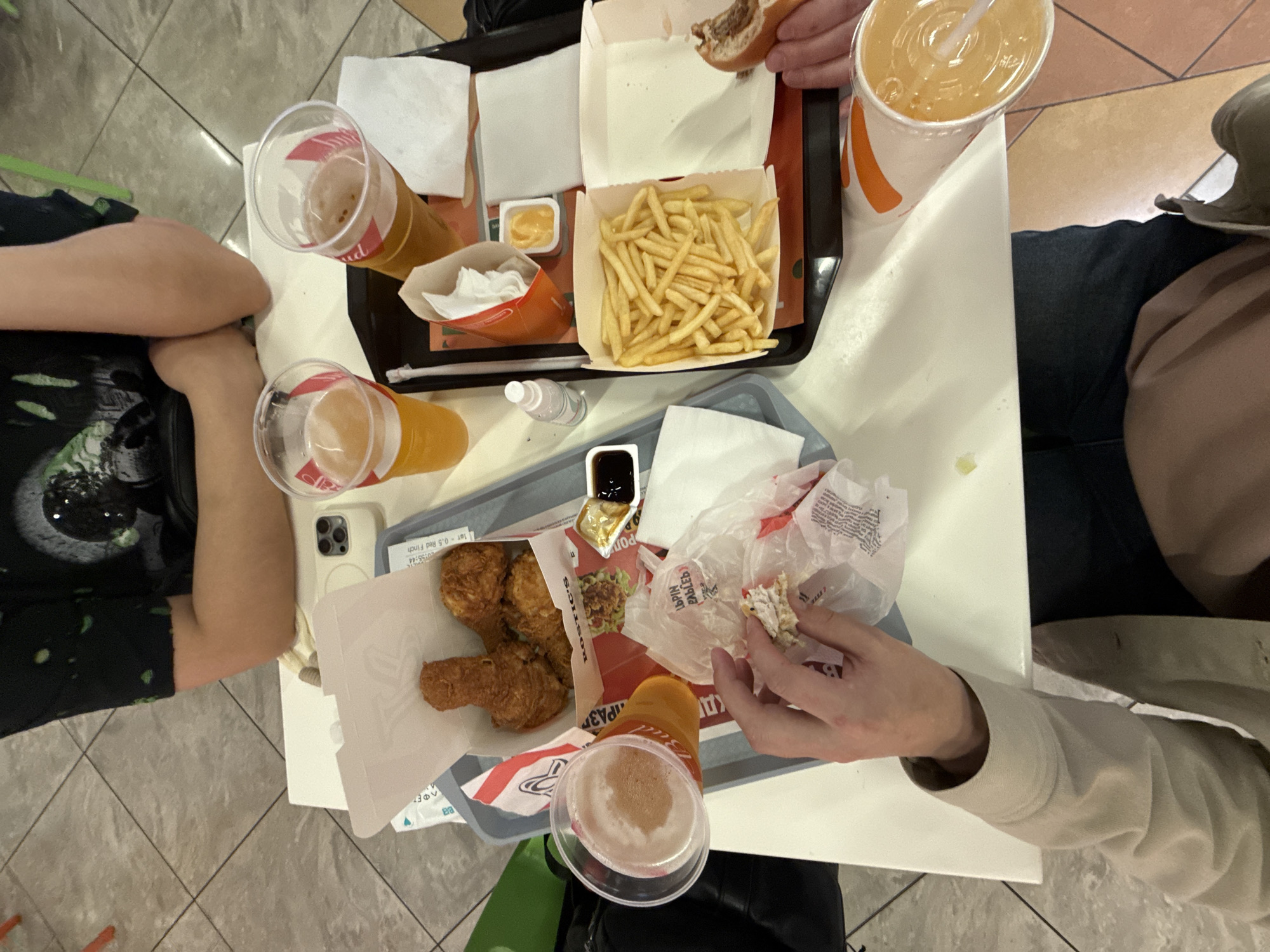 The image shows a table with food and drinks, likely in a restaurant. Two people are visible, one wearing a dark T-shirt with a graphic design and another wearing a light-colored jacket. On the table, there is a tray of fast food including french fries, a burger, a container of sauce, fried chicken pieces, and drinks in plastic cups. The location is indicated as Moscow, Russia, based on the Cyrillic writing on the chicken packaging. One person is seen holding and eating food.