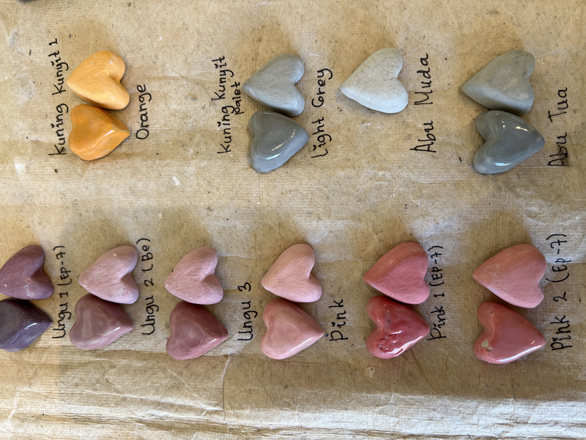 The image displays an overhead view of various heart-shaped material samples arranged in columns on a light brown, textured paper surface, suggestive of a color or material palette. The samples demonstrate different colors and finishes.
On the left side, six pairs of heart samples are arranged vertically. The top pair consists of one dark purple glossy heart and one matte mauve heart, labeled "Ungu 1 (Ep-7)". Below this, a pair of medium mauve hearts, one glossy and one matte, are labeled "Ungu 2 (Be)". The next pair, featuring one glossy and one matte light dusty pink heart, is labeled "Ungu 3". Following this is a pair of light pink hearts, one glossy and one matte, simply labeled "Pink". The fifth pair consists of glossy and matte medium red-pink hearts, labeled "Pink 1 (Ep-7)". The bottom-most pair on the left features glossy and matte deep red-pink hearts, labeled "Pink 2 (Ep-7)".
On the right side, there are three distinct sets of grey and orange samples. The top pair consists of one glossy bright orange heart and one matte slightly paler orange heart, labeled "Kuning Kunyit 1" and "Orange". Below this, two light grey hearts, one glossy and one matte, are labeled "Kuning Kunyit Palet" and "Light Grey". A single off-white grey heart with a cracked texture sits beneath this pair, labeled "Abu Muda". The lowest set on the right features two darker grey hearts, one glossy and one matte, labeled "Abu Tua".
The background paper exhibits creases and scattered small white specks. The overall scene suggests a collection of handcrafted or artistic samples. This scene is located in Ubud District, Indonesia.