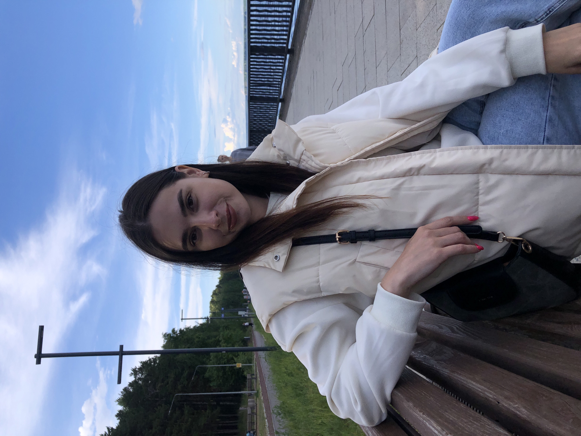 The image shows a woman sitting on a park bench. She has long dark hair, light skin, and is wearing a white jacket over a white hoodie and light blue jeans. She is holding a black handbag with a black strap. The background consists of trees, grass, street lights, and a metal railing that suggests a waterfront or elevated area. The sky is visible in the background with some clouds. The location may be a park or promenade near a body of water in Chaikovsky, Russia, although specific landmarks confirming the exact location are not visible in the photograph.