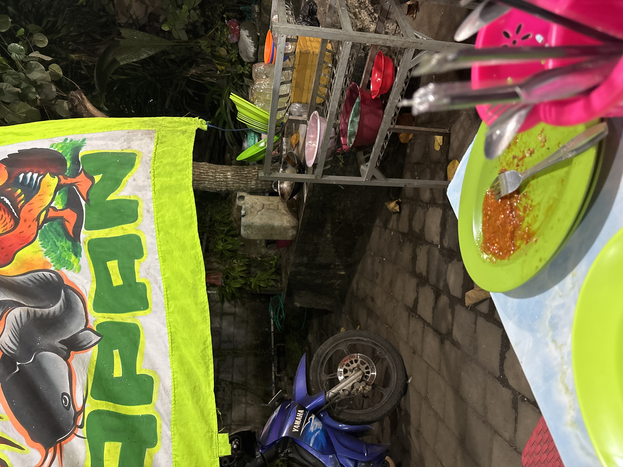 This image depicts an outdoor food stall setting in Kuta Selatan, Indonesia. A blue Yamaha motorcycle is parked to the left, partially obscuring a wall. A tarp with cartoonish drawings of a catfish and a rooster, along with green lettering, is strung up above. In the foreground, a light blue table holds two bright green plates. One plate contains red chili sauce and a fork. A bright pink utensil holder filled with silverware is to the right of this plate. Behind the table, a metal shelf unit holds various bowls and glasses. The ground is paved with interlocking grey bricks. The background features lush green foliage and trees, suggesting a tropical environment. The lighting suggests it is either dusk or nighttime.