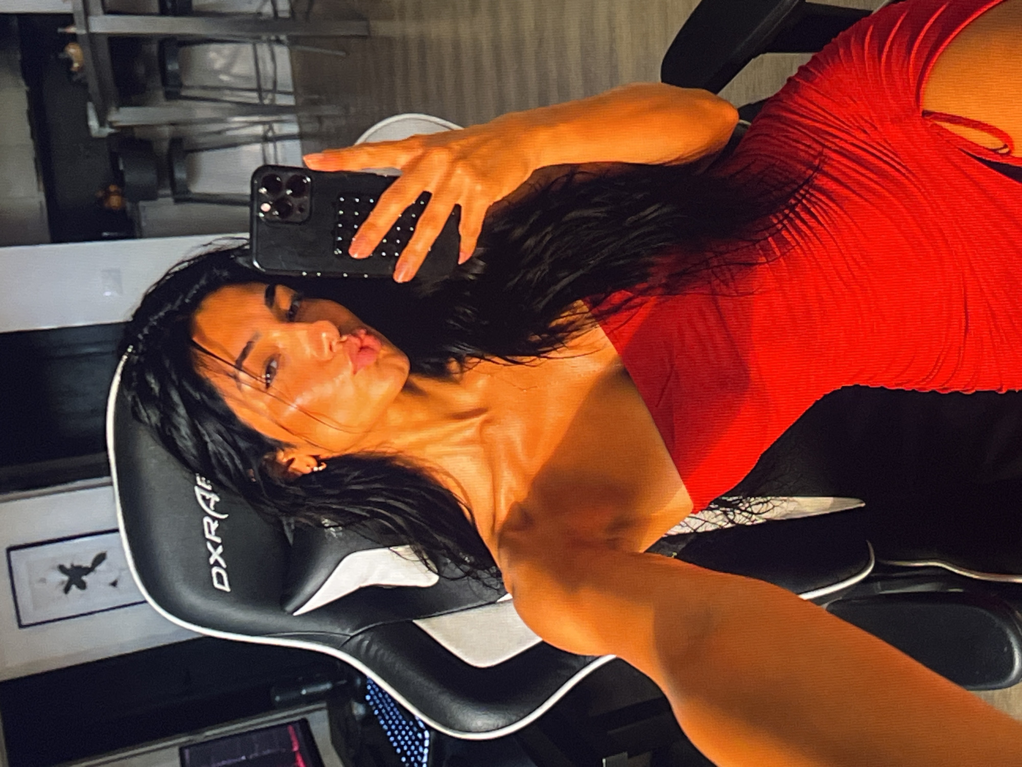 A young woman with long, dark hair is taking a selfie in a room. She is wearing a strapless red dress with ruching and a slit on the side. She is seated in a black and white gaming chair with the "DXRacer" logo visible. Her right arm is extended, holding a black smartphone to take the picture. Her lips are pursed. In the background, a framed picture hangs on the wall, and a blurry kitchen area with stools and a counter is visible. There is also a computer monitor and keyboard to the left. The lighting in the room casts a warm, orangish glow on the woman.