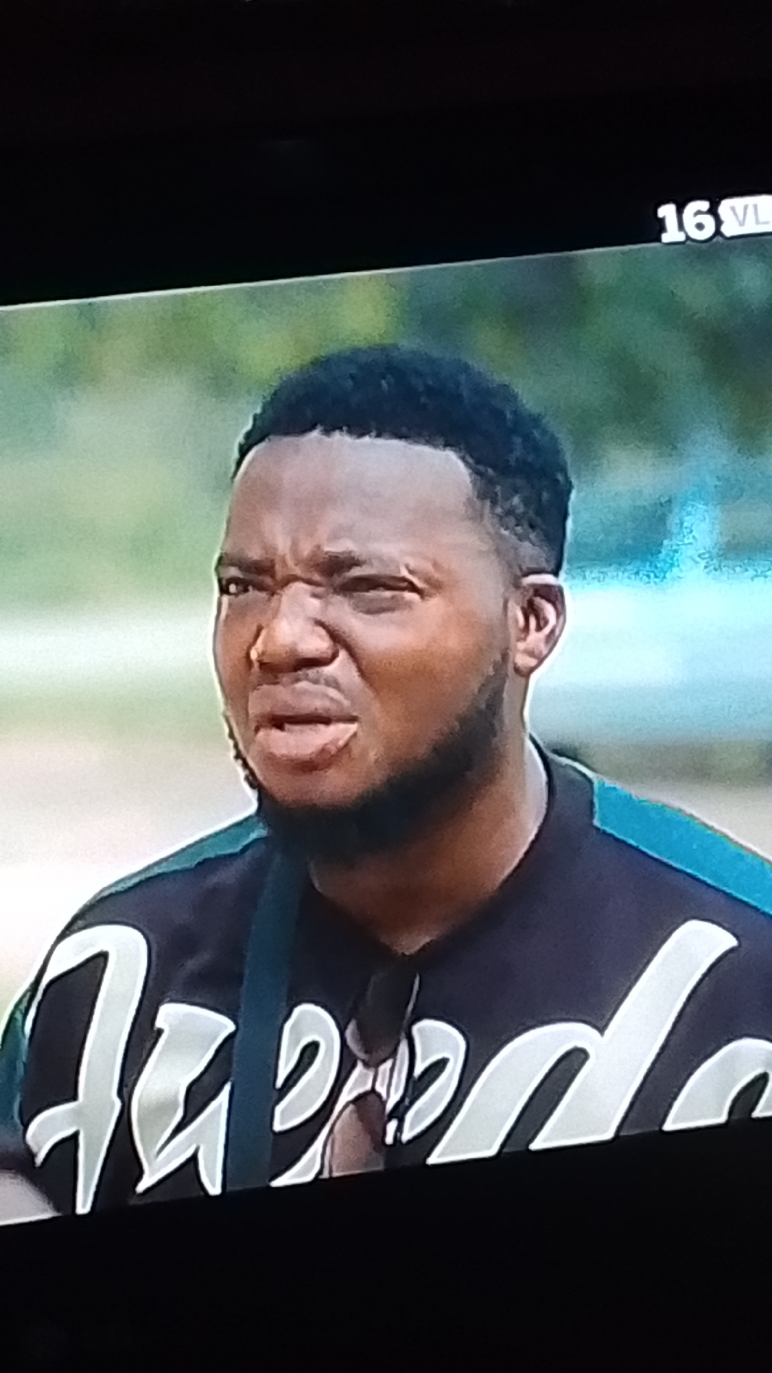 A forensic analysis of the provided image reveals a close-up still from a video sequence, ostensibly set in the city of Lokoja, Nigeria. The primary subject is a dark-skinned adult male, positioned centrally, with a short, dark haircut featuring a faded side and a neatly trimmed beard. His facial expression displays significant emotional intensity, characterized by furrowed brows, squinted eyes, and a slightly open mouth, indicating a state of discomfort, disgust, or deep concentration. He is wearing a dark-colored polo shirt, possibly black or dark blue, with stylized light-colored lettering on the front, partially visible as "Agedo". A teal or light blue stripe is evident on the upper right shoulder of the shirt. A dark strap, potentially from a bag or equipment, crosses over his right shoulder. The background is an out-of-focus expanse of green foliage, suggesting an outdoor environment. In the upper right corner of the frame, a white alphanumeric overlay displays "16 VL", likely denoting a content rating.