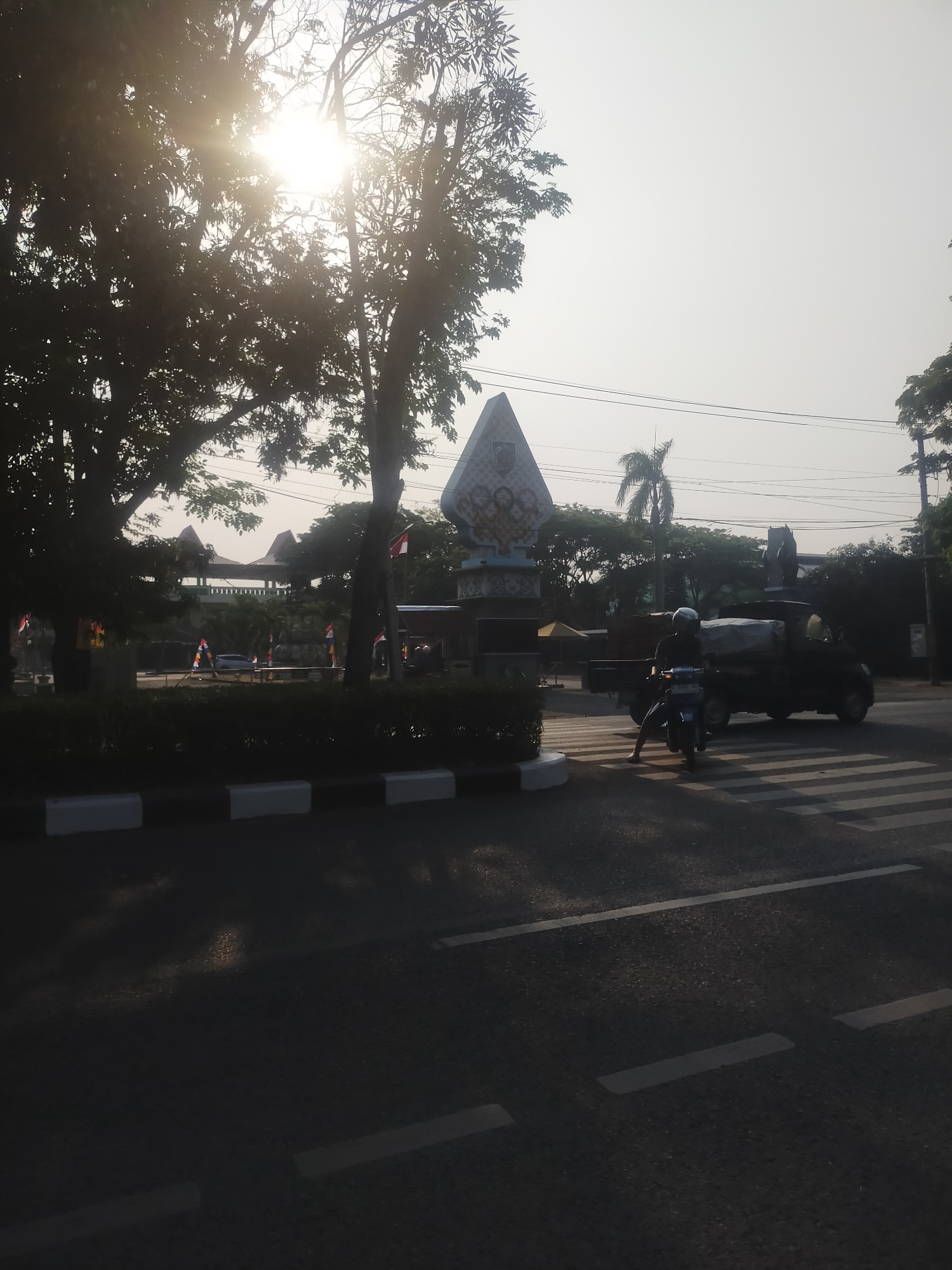 The image captures a street scene in Pedagangan, Indonesia, during the morning as indicated by the low-angled sunlight filtering through trees. In the foreground, a crosswalk is visible on the asphalt road. To the left, a person on a scooter is preparing to cross, with a small truck laden with goods behind them. In the background, a large triangular monument stands prominently, adorned with intricate carvings and possibly a crest. Flags, including the Indonesian flag, are displayed on poles, suggesting a public or commemorative area. Trees and other vegetation line the street, and in the distance, a traditional Indonesian-style pavilion or structure can be seen. The sky is hazy, characteristic of some tropical urban environments.