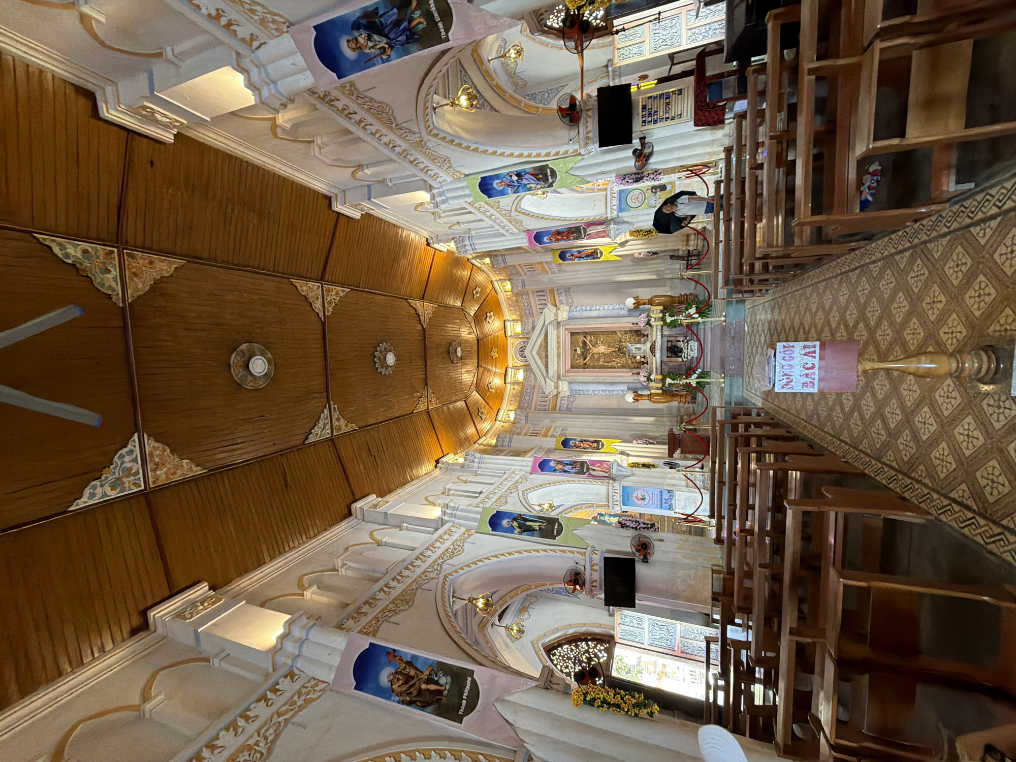 The image shows the interior of a church in Hội Tín, Vietnam. The church has a wooden ceiling with decorative elements and white walls adorned with religious banners. Pews are arranged in rows, leading towards an altar at the front of the church. The altar features a crucifix and floral decorations. There are various religious statues and artwork visible throughout the church. A young person holding a black umbrella stands near the altar area. The flooring is tiled with a patterned design.