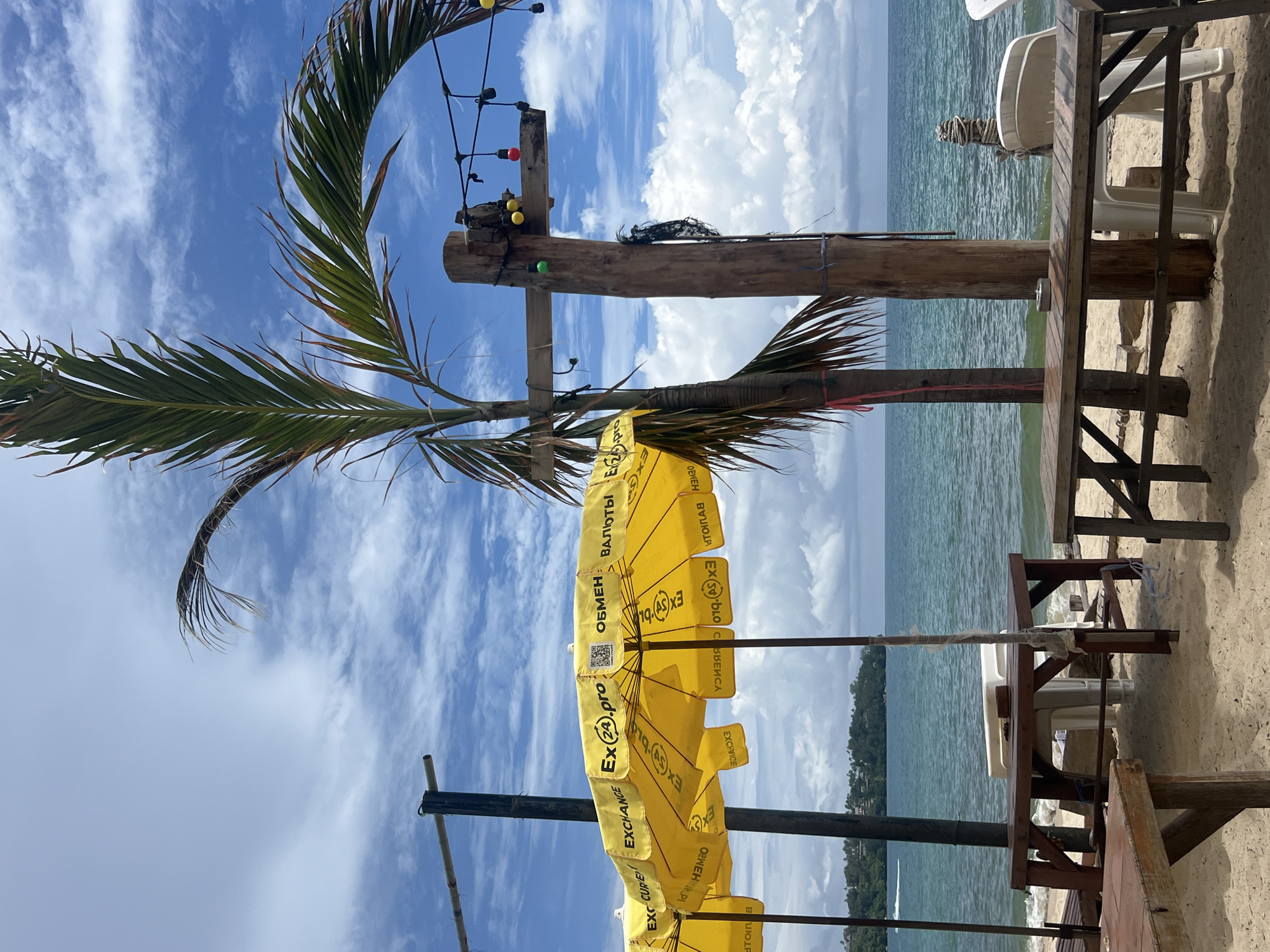 This is a photograph of a beach in Choeng Thale, Thailand, on a bright, cloudy day. The foreground is dominated by a sandy beach with several wooden tables and chairs, suggesting a seating area for a beachside establishment. Two yellow beach umbrellas with the branding "EX24.pro" and Cyrillic text are prominently displayed, one partially obscuring the other.
A tall palm tree with its fronds reaching towards the sky is positioned to the right of the umbrellas. Next to the palm tree, a rustic wooden pole serves as a support structure, adorned with a string of colorful decorative lights—red, yellow, and green bulbs—and a piece of black netting.
In the background, the turquoise ocean stretches out under a sky filled with white, fluffy clouds. The water appears calm, with gentle waves lapping at the shore. Beyond the water, a lush green coastline with trees and some buildings is visible, indicating the presence of land and possibly a town or resort area.
The overall scene conveys a relaxed, tropical atmosphere, typical of a beachfront location in Thailand.