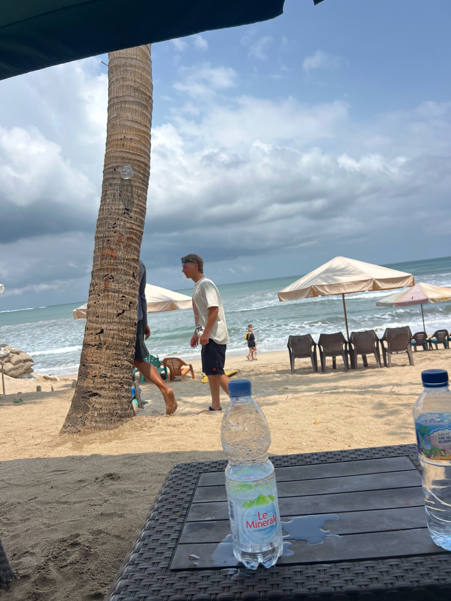 The image depicts a beach scene in Kuta, Indonesia. A large palm tree stands prominently in the foreground, partially obscuring two individuals walking on the sand. The ocean with moderate waves is visible in the background, with a cloudy sky overhead. Several beach chairs and umbrellas are set up on the sand. In the immediate foreground, a woven table holds two bottles of water. One bottle is labeled "Le Minerale," and the other has a label with text and imagery suggesting it contains a beverage. The overall environment suggests a relaxed beach setting, likely a resort or public beach area in Kuta.