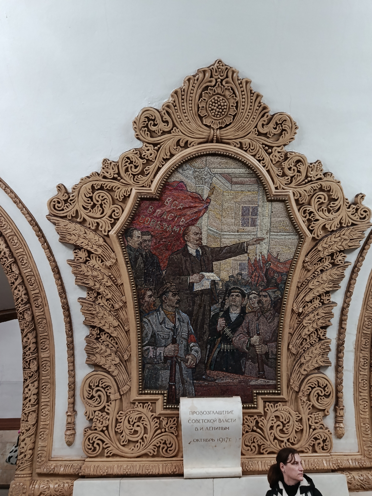 The image shows a mosaic depicting Vladimir Lenin addressing a crowd, set within an ornate, carved frame with floral and wheat motifs. The mosaic includes a red banner with Cyrillic script and depicts armed people. Below the mosaic is a plaque written in Cyrillic, which translates to "Proclamation of Soviet Power by V.I. Lenin, October 1917".
The scene appears to be located in a Moscow metro station due to the curved tunnel wall with similar decorations on the left side. A person can be seen in the lower right corner of the image.
