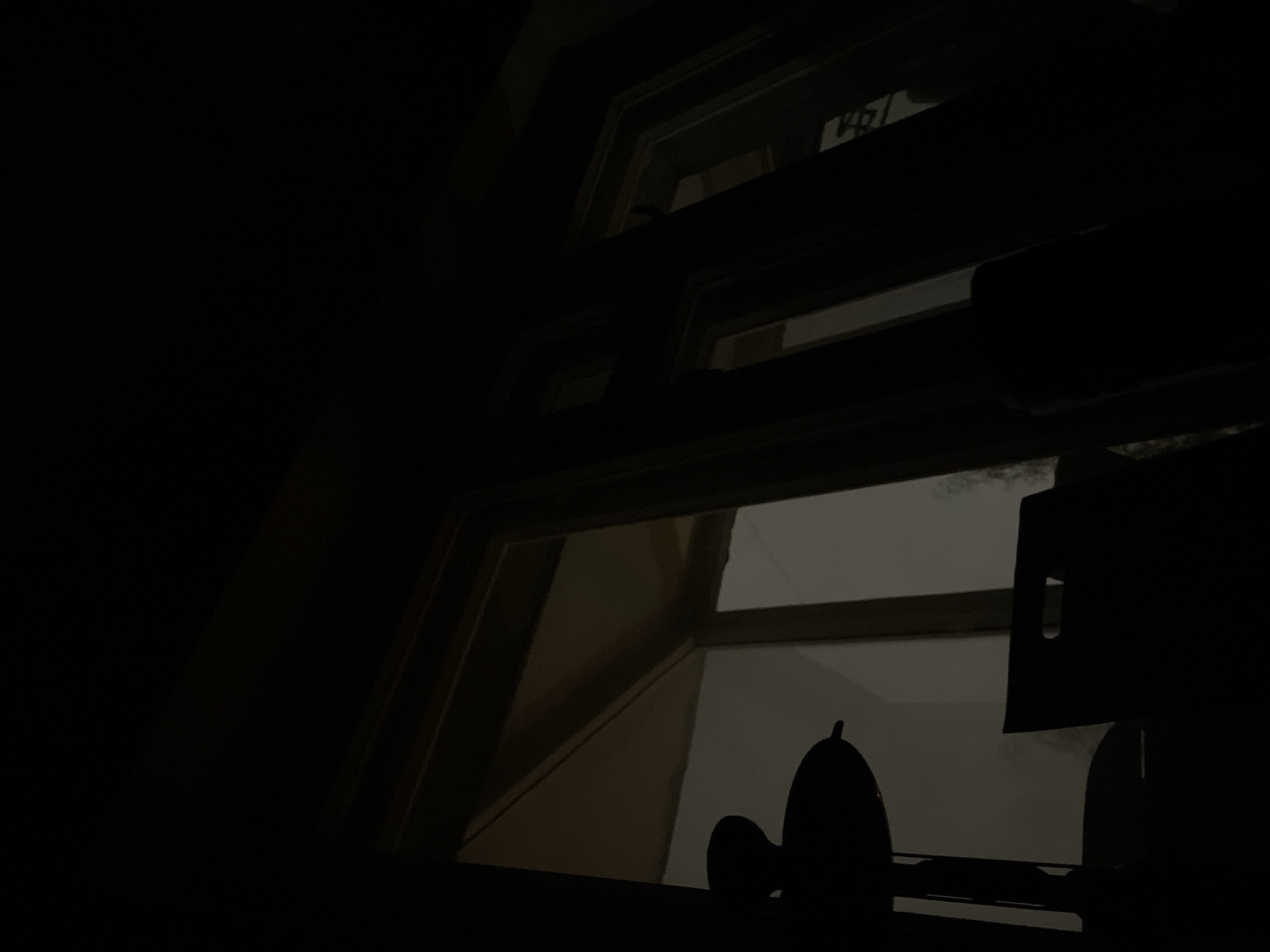 A low-angle, extremely dark indoor shot looking out of a window. The window frame is white and segmented. Outside, there is a blurry, indistinct view, likely of an overcast or night scene. Silhouetted against the window is an unidentifiable object, possibly a decorative item. No people are visible. There's no information suggesting Murino, Russia.
