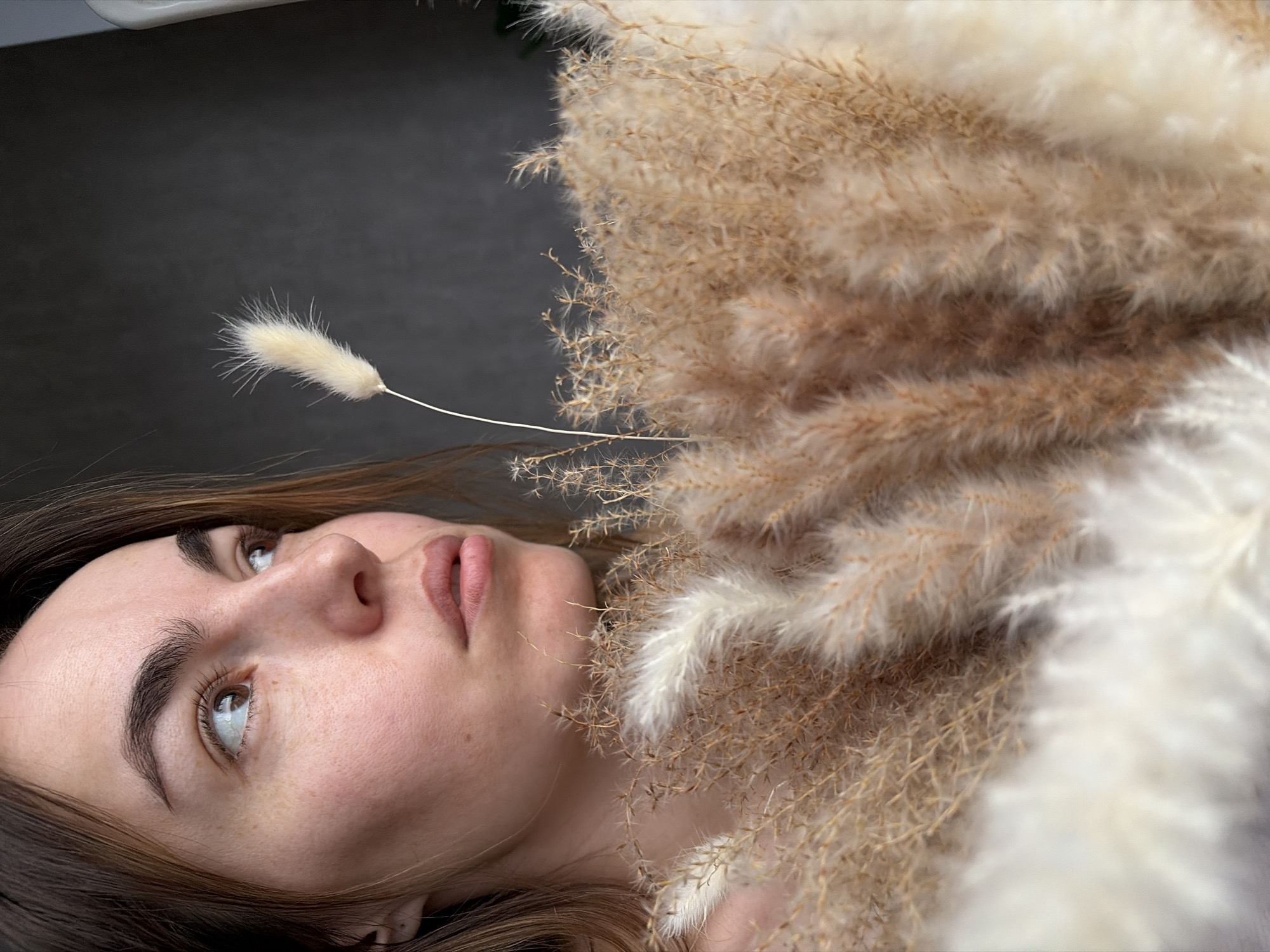 A woman lies with dried floral arrangements, likely indoors with a dark grey background. The floral arrangements feature various textures and colors, ranging from white to beige and brown. The woman has fair skin, brown eyes, and dark eyebrows. She has freckles and is looking upwards. The background is a dark grey. There is no indication of Izhevsk, Russia, or any identifiable city location in the image.