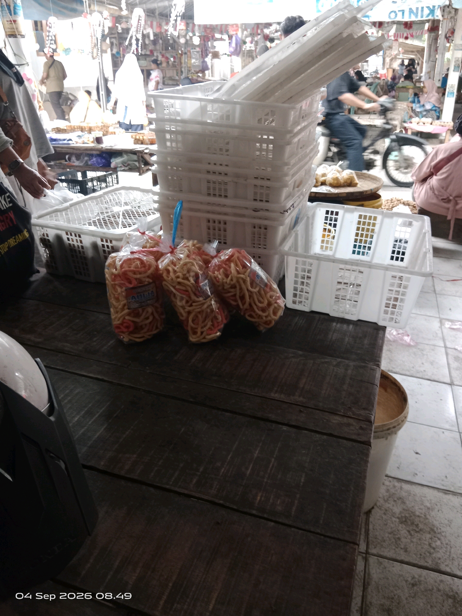 In the city of Kalipurwo, Indonesia, a market scene unfolds. In the foreground, a dark wooden table dominates the frame, its surface weathered and showing distinct wood grain. To the left of the table, a black object, possibly a scooter's front panel, is visible. Resting on the table are three plastic bags filled with coiled snacks, each tied with a red string, and a blue plastic scoop.
Behind the table, a stack of white plastic crates is arranged. To the right of these crates, another white plastic crate is positioned, with an empty bucket partially visible beneath it. Further in the background, the market environment is bustling. Several people are present, some blurred due to motion. A man is riding a motorcycle, and others are engaged in market activities. The stalls display various goods, with a bright, sunny atmosphere suggesting an outdoor or semi-enclosed market setting. The overall impression is one of a typical Indonesian market day.