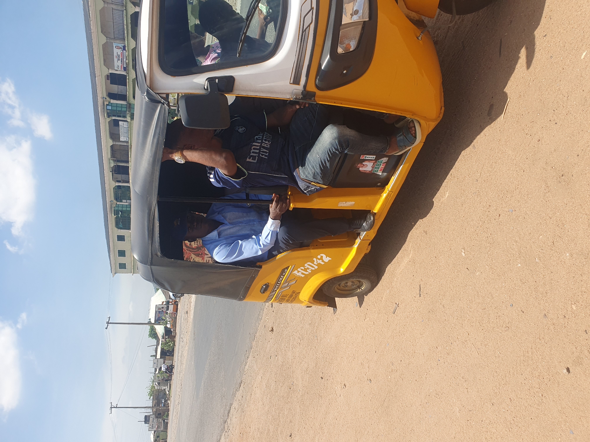 This daytime image, captured in Lokoja, Nigeria, features a yellow and white three-wheeled auto-rickshaw, commonly known as a Keke Napep, positioned on a dirt road beside an asphalt street. The vehicle appears to be stationary or moving slowly.
Inside the auto-rickshaw, at least three men are visible. The driver, wearing a light blue long-sleeved shirt and dark trousers, is seated at the controls. Behind him, a passenger in a dark blue t-shirt, emblazoned with "Emirates Fly Better," and blue jeans, is leaning back. A watch is visible on his left wrist. Another passenger in a light blue shirt is partially seen in the rear, while a person in a pink shirt is visible in the reflection of the front windshield.
The side of the yellow vehicle bears the registration number "FC042" in black, along with "DELUXE PLUS" and smaller text that includes "DELHI AUTO" and "SHAKTI." A "BABA" sticker is present inside the passenger area.
The setting includes a dusty, light brown dirt road bordering a darker asphalt road. In the background, multi-story buildings with light-colored walls and green or blue-tinted windows line the street. One building displays a banner with the word "PROMISE" and other obscured text, suggesting commercial activity. Utility poles with overhead wires are spaced along the road. The sky is bright blue with scattered white clouds, indicating clear, sunny weather. The scene depicts a common street environment in a Nigerian town.