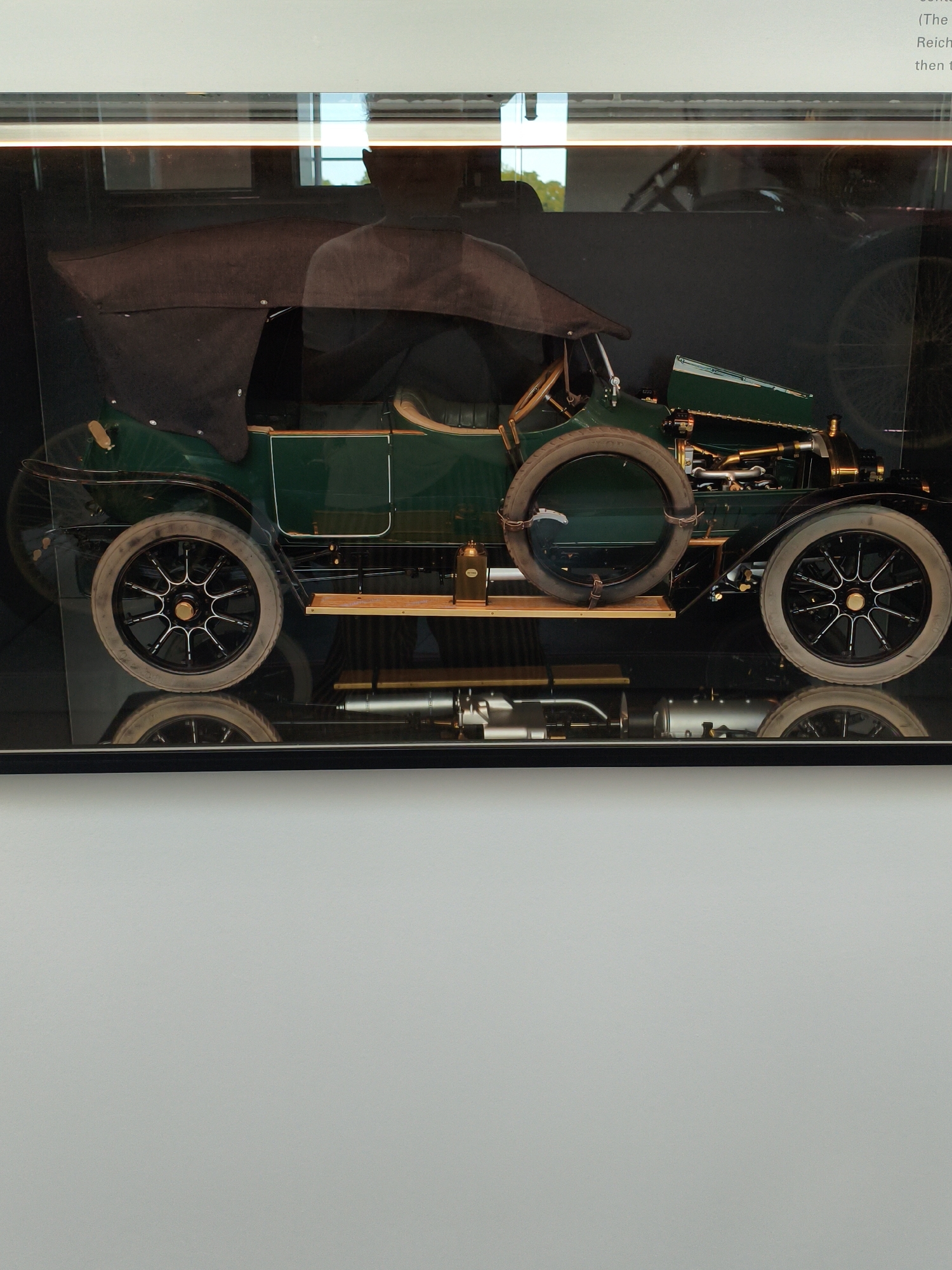 The image displays an exhibit inside a glass case. The primary subject is an antique green car with a dark brown convertible top. The car has spoked wheels with white rims and black tires. The interior of the car is visible, including a wooden steering wheel and upholstered seats. In the foreground, there is a reflection of the car, appearing on a dark, polished surface. Behind the car, a large spoked wheel is visible. Text is present at the top right of the image, indicating the context of the exhibit, though the text is partially obscured and illegible in detail. The exhibit appears to be located in a museum or exhibition space. Based on external information, this image was taken at the Audi Forum in Ingolstadt, Germany.