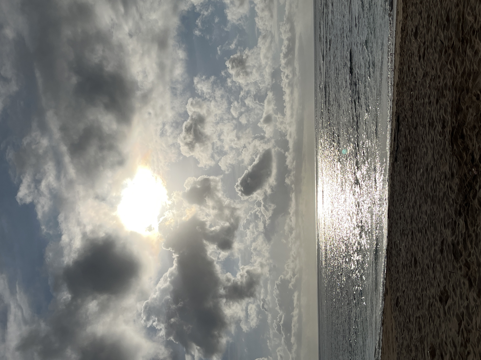 This image depicts a scene at a beach in Kuta Selatan, Indonesia. The view is horizontal with the sky occupying the left side and the beach and sea on the right. The sky is filled with clouds, and the sun is visible through some of them, causing a bright glare. The sea reflects the sunlight, creating a shimmering effect on the water's surface. The sand on the beach appears dark and grainy, indicating a possible coarse texture. There is no visible evidence of people, subjects, or actions in the photograph.