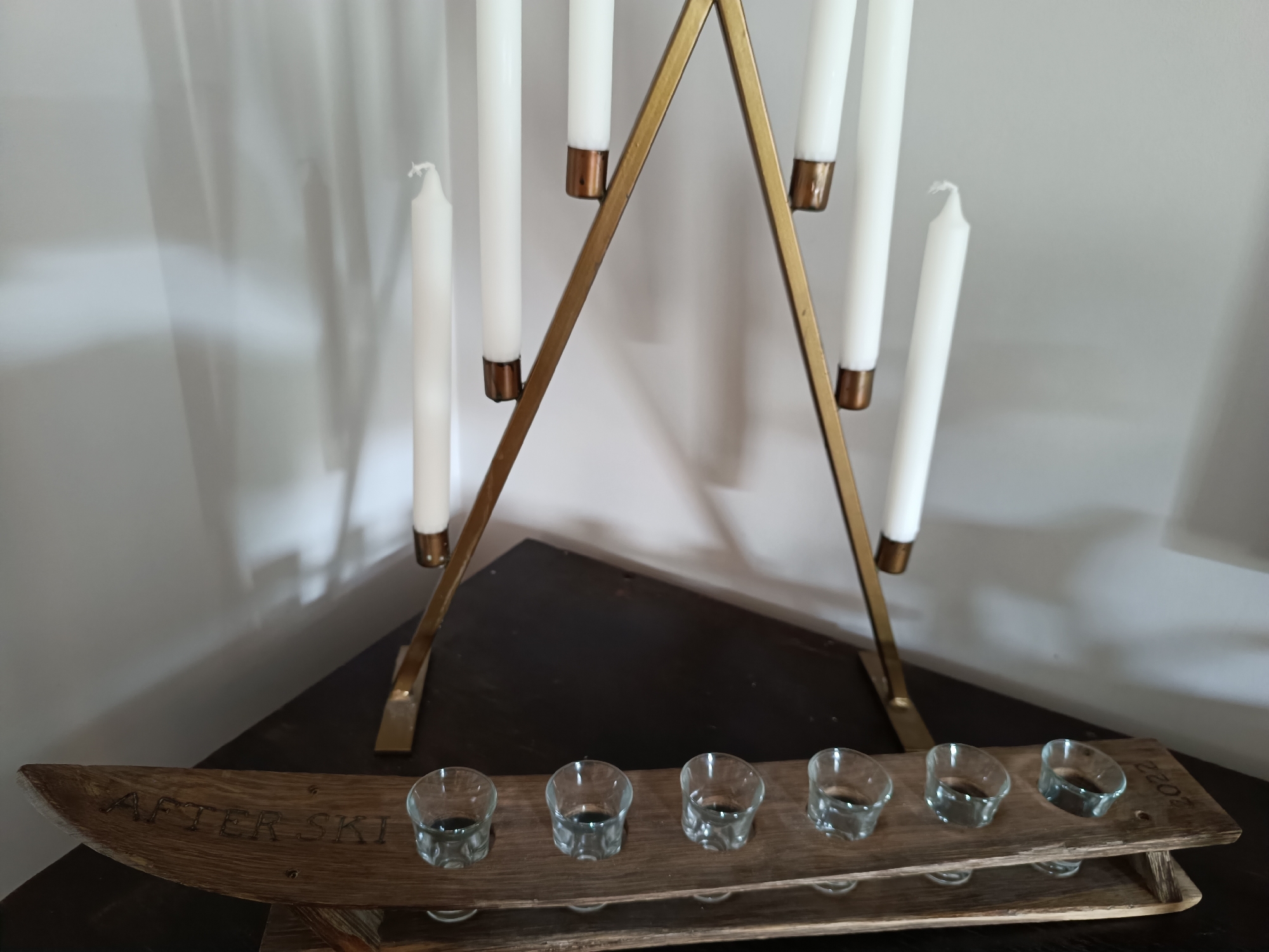 In an indoor setting, likely a home in Hovden, Norway, a wooden display with the text "AFTER SKI 2022" is visible, holding seven small shot glasses. Above the display, a gold-colored candelabra holds seven white taper candles. The scene is lit by ambient light, casting shadows on a white wall in the background.